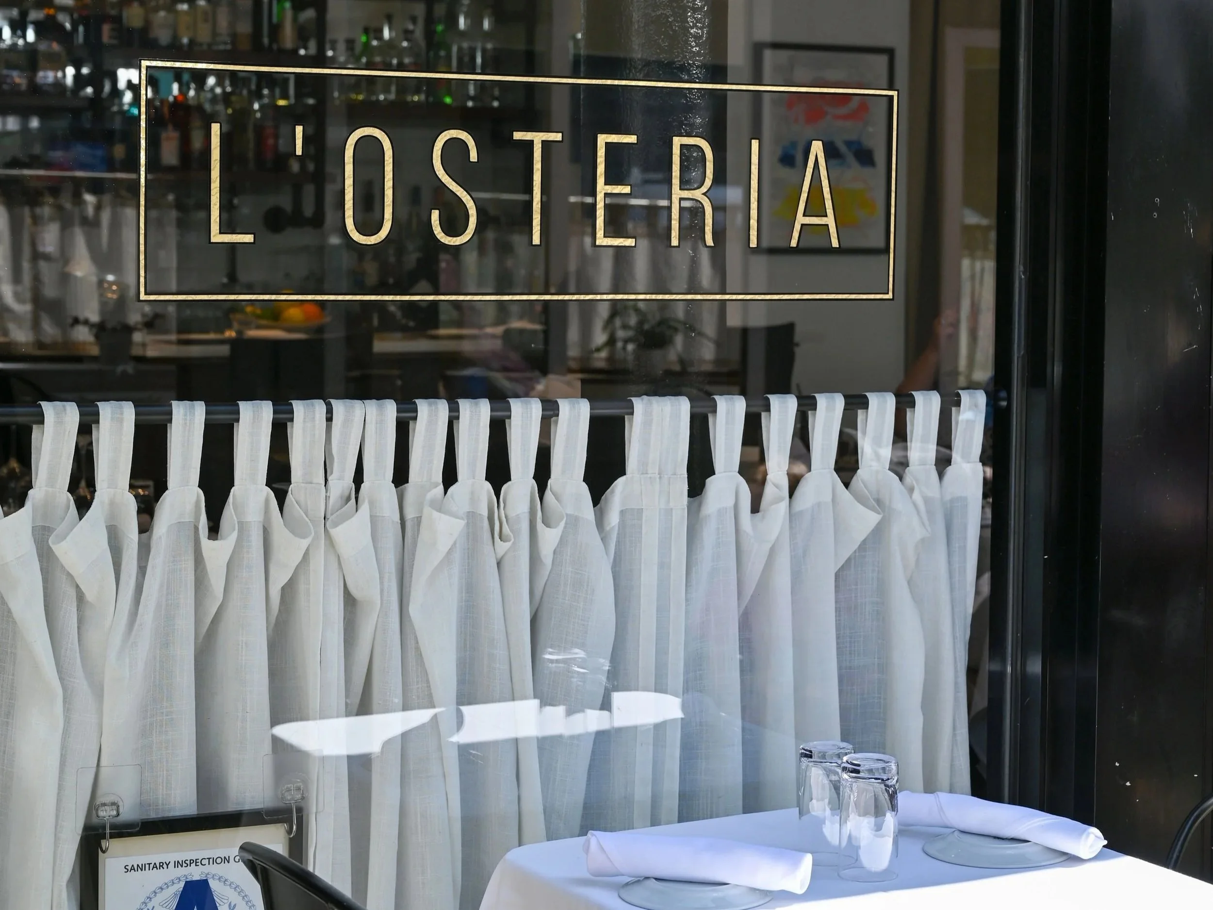 A restaurant or cafe table set with a white tablecloth, two upside-down glasses, and a napkin with utensils, behind a window with a curtain. The window has a sign that says 'L'OSTERIA' and a label that reads 'SANITARY INSPECTION'.