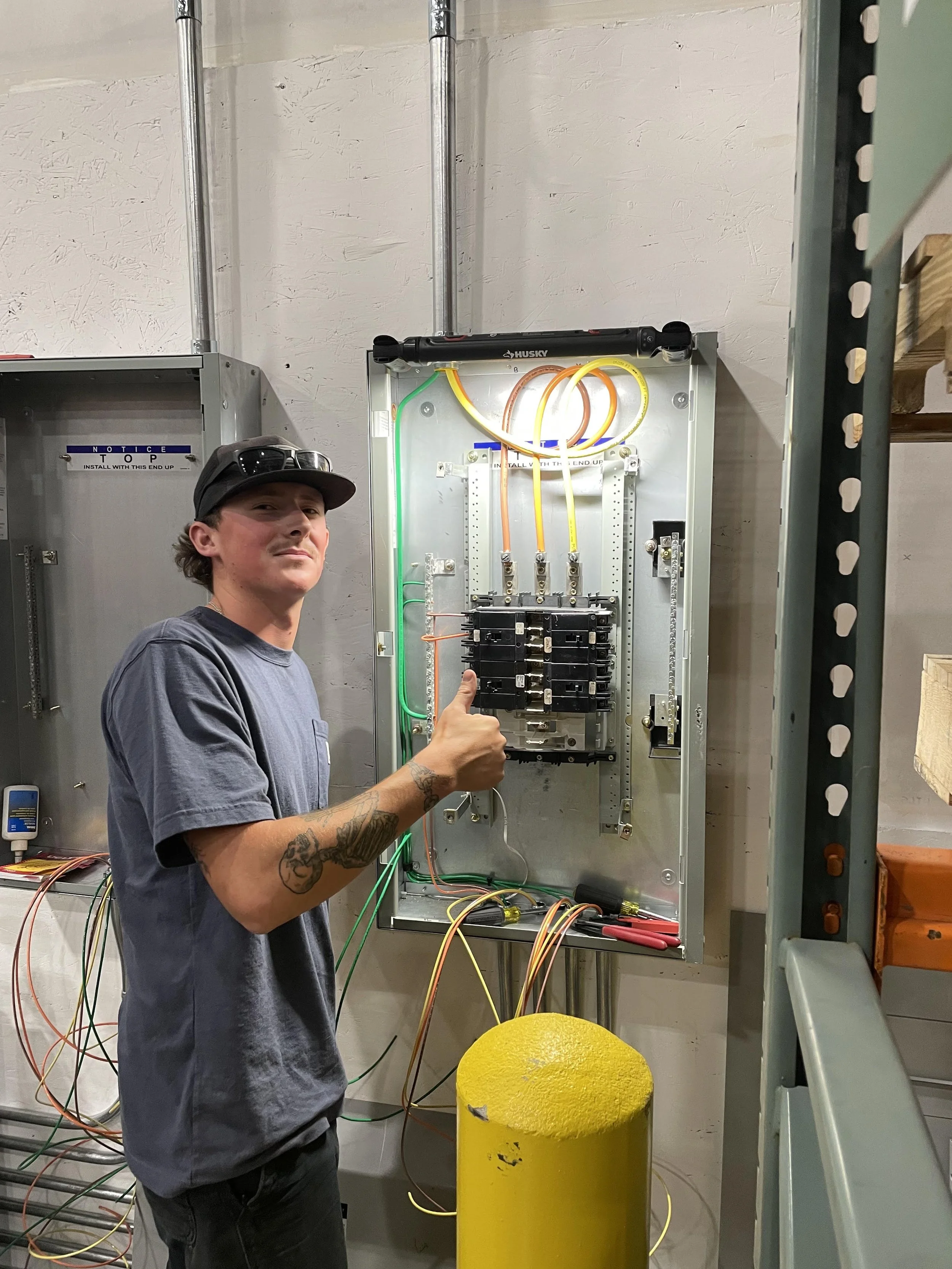 A young man with tattoos on his arm, wearing a gray t-shirt, black cap, and sunglasses on his head, giving a thumbs up while working on an electrical panel with wires. The panel contains brightly colored wires and circuit breakers, situated in an industrial or workshop setting.