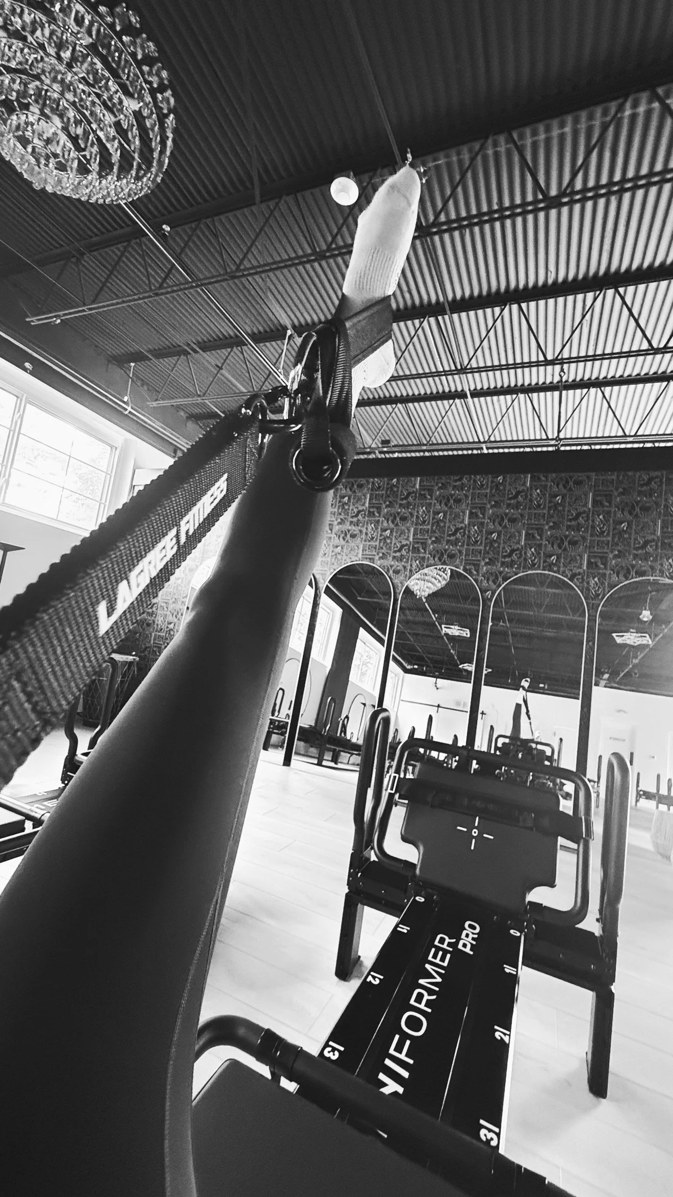 Black-and-white view along a Lagree machine, a foot in a Lagree Fitness strap reaching toward a crystal chandelier