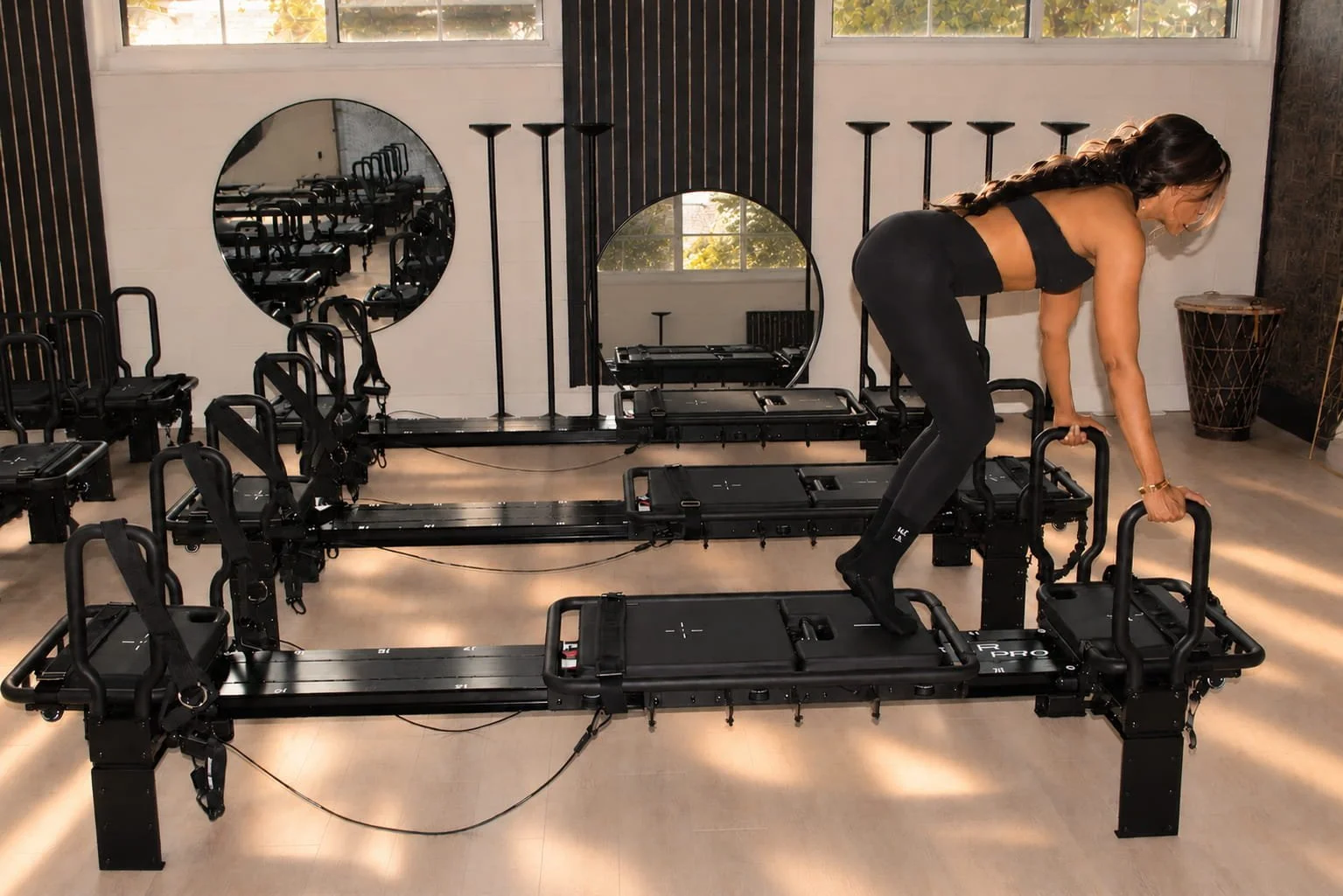 A woman steps onto the carriage of a Lagree machine, hands on the front handles, in a sunlit studio