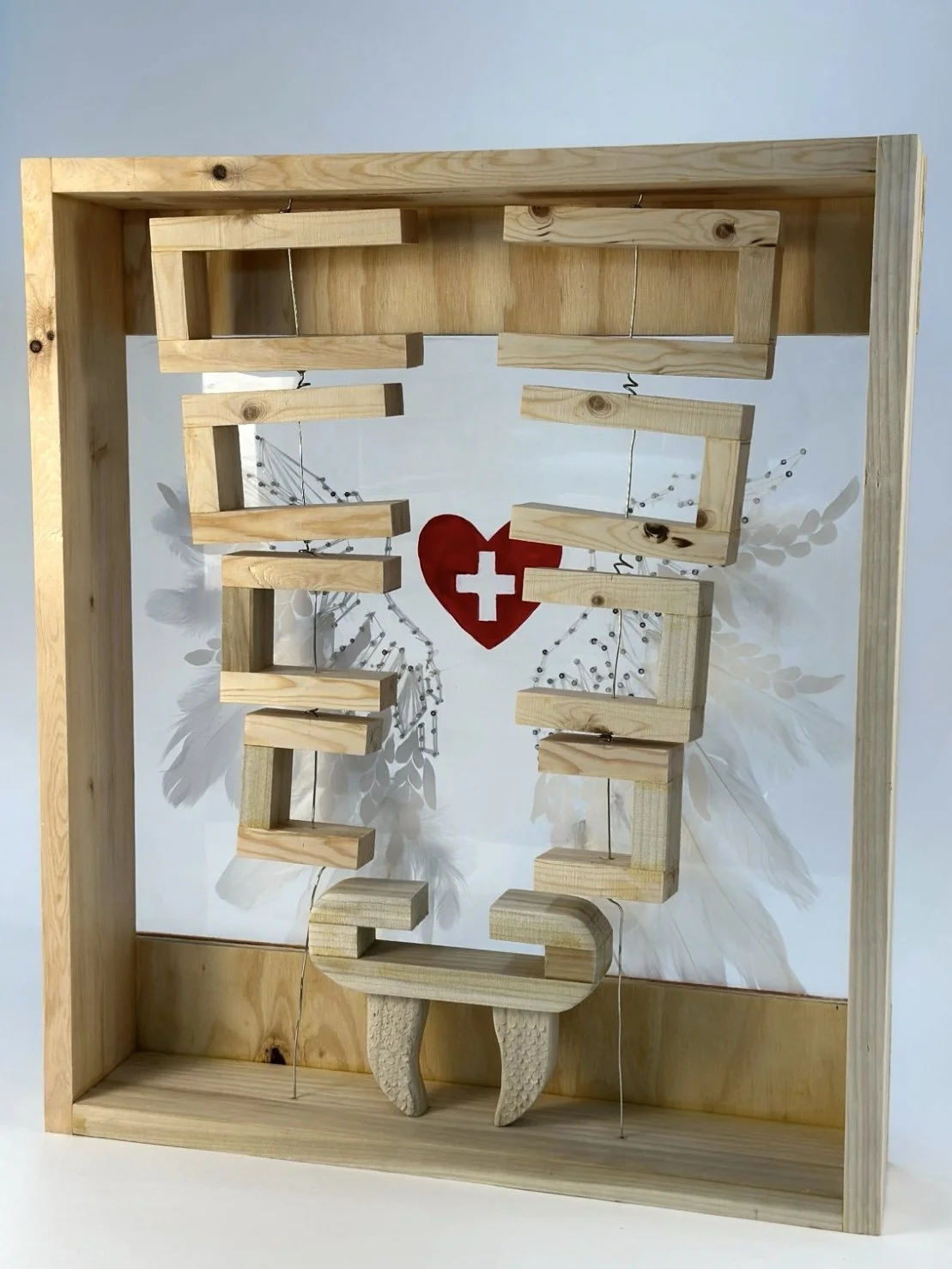 The sculpture combined wood and transparent acrylic sheets. The wooden box represents a first-aid kit with abstract symbols of two snakes, angel wings, and a red heart.