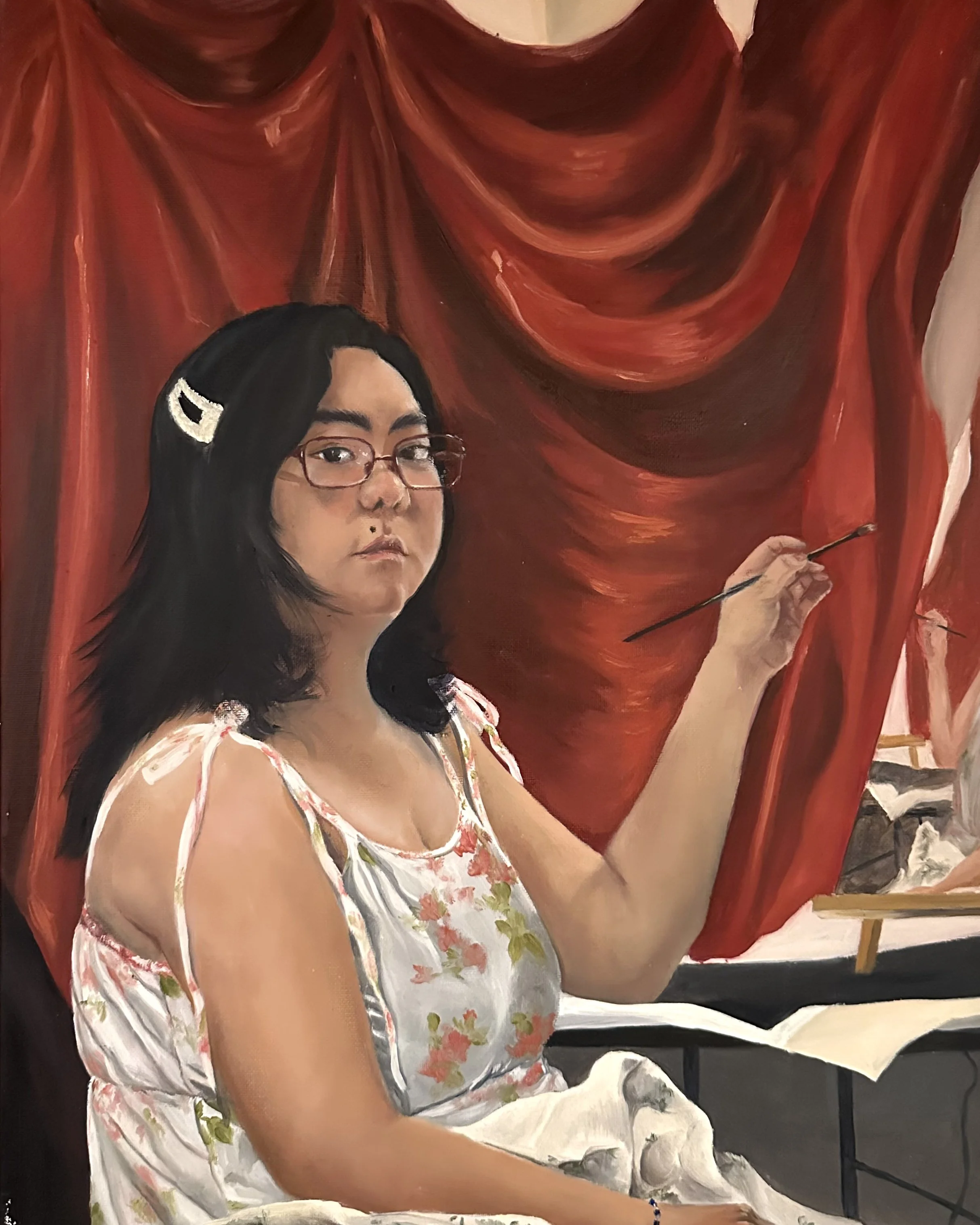 A woman with black hair and glasses is painting on a large canvas featuring red draped fabric and a portrait of herself. The woman is dressed in a floral dress and is holding a paintbrush.