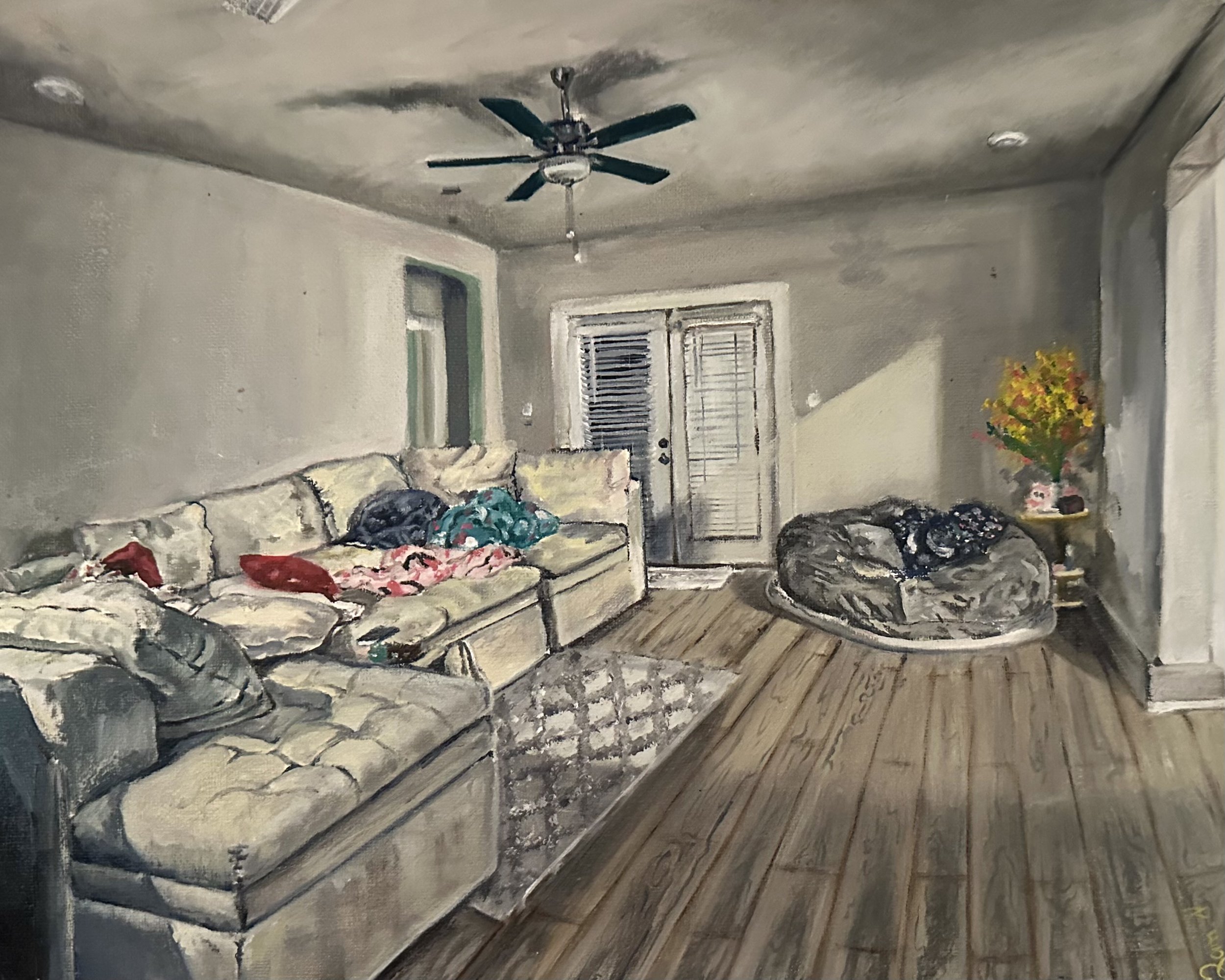 Living room with beige couch, colorful pillows, ceiling fan, large window with blinds, door, and a flower arrangement on a side table.