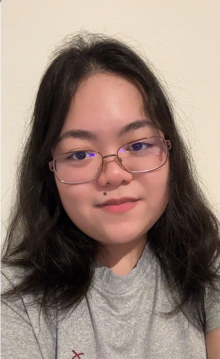 A young woman with black hair, glasses, and a mole above her lip, wearing a gray shirt, smiling at the camera against a plain, light-colored wall.