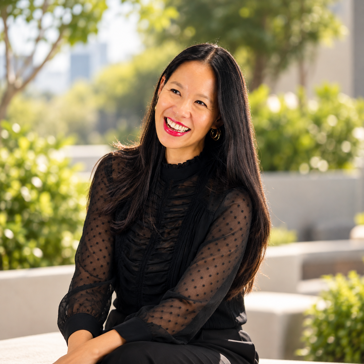 A woman with long black hair and bright red lipstick smiling outdoors in front of greenery and skyscrapers.