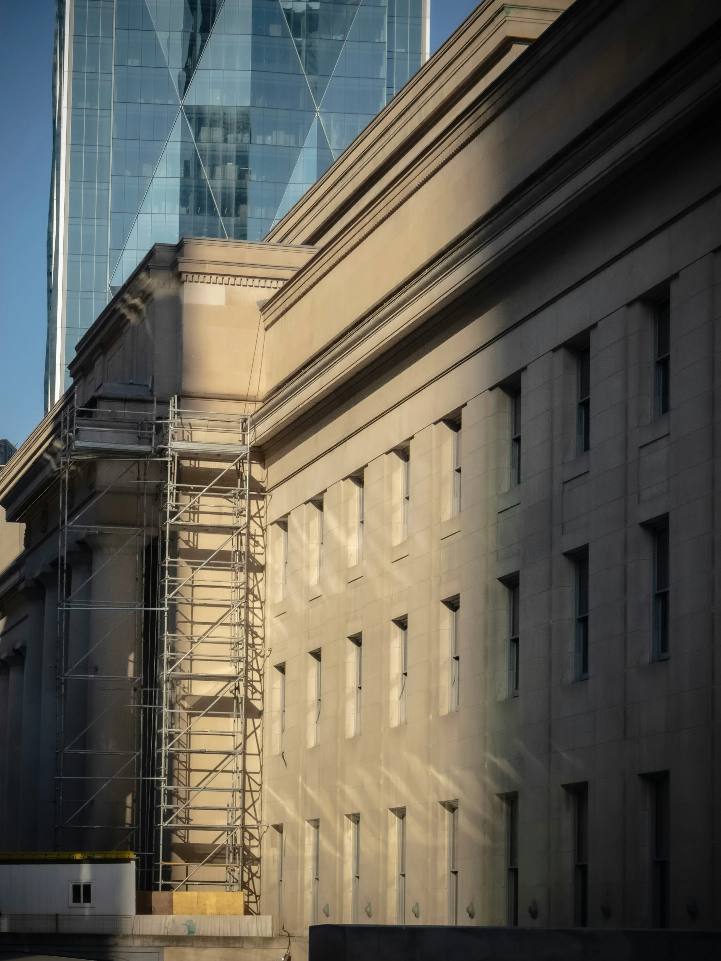 A modern city building with a glass exterior reflecting the sky, adjacent to an older building with classical architecture and scaffolding at the corner.