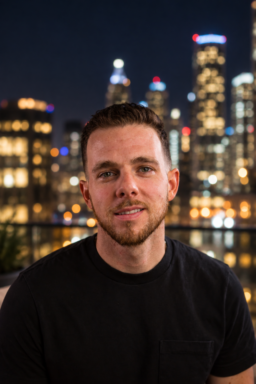 A young man with short brown hair and a beard, wearing a black T-shirt, sitting outdoors at night with a city skyline and lit-up skyscrapers in the background.