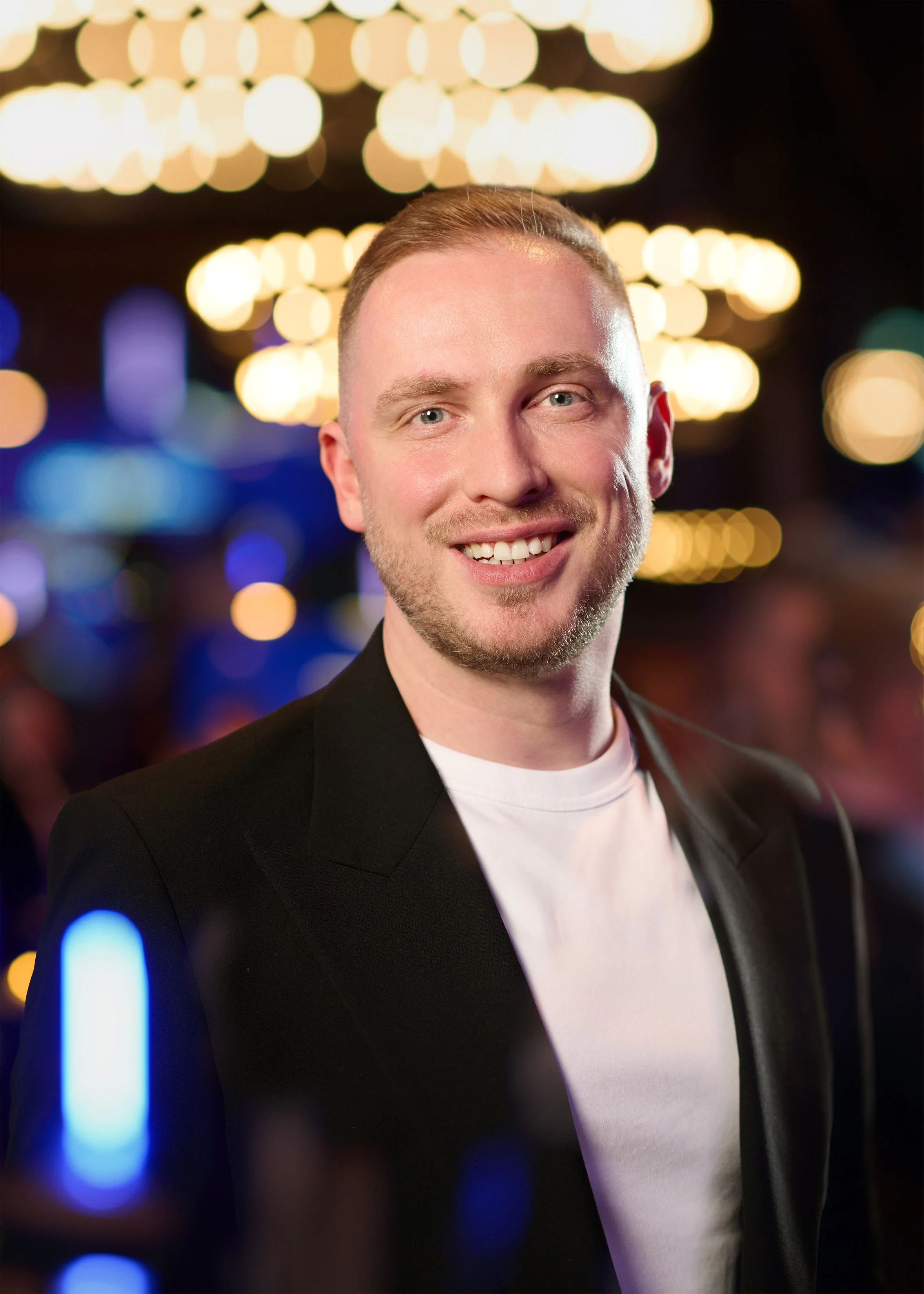 A young man with short light brown hair and blue eyes smiling, wearing a black blazer over a white shirt, with city lights and bokeh effect in the background at night.