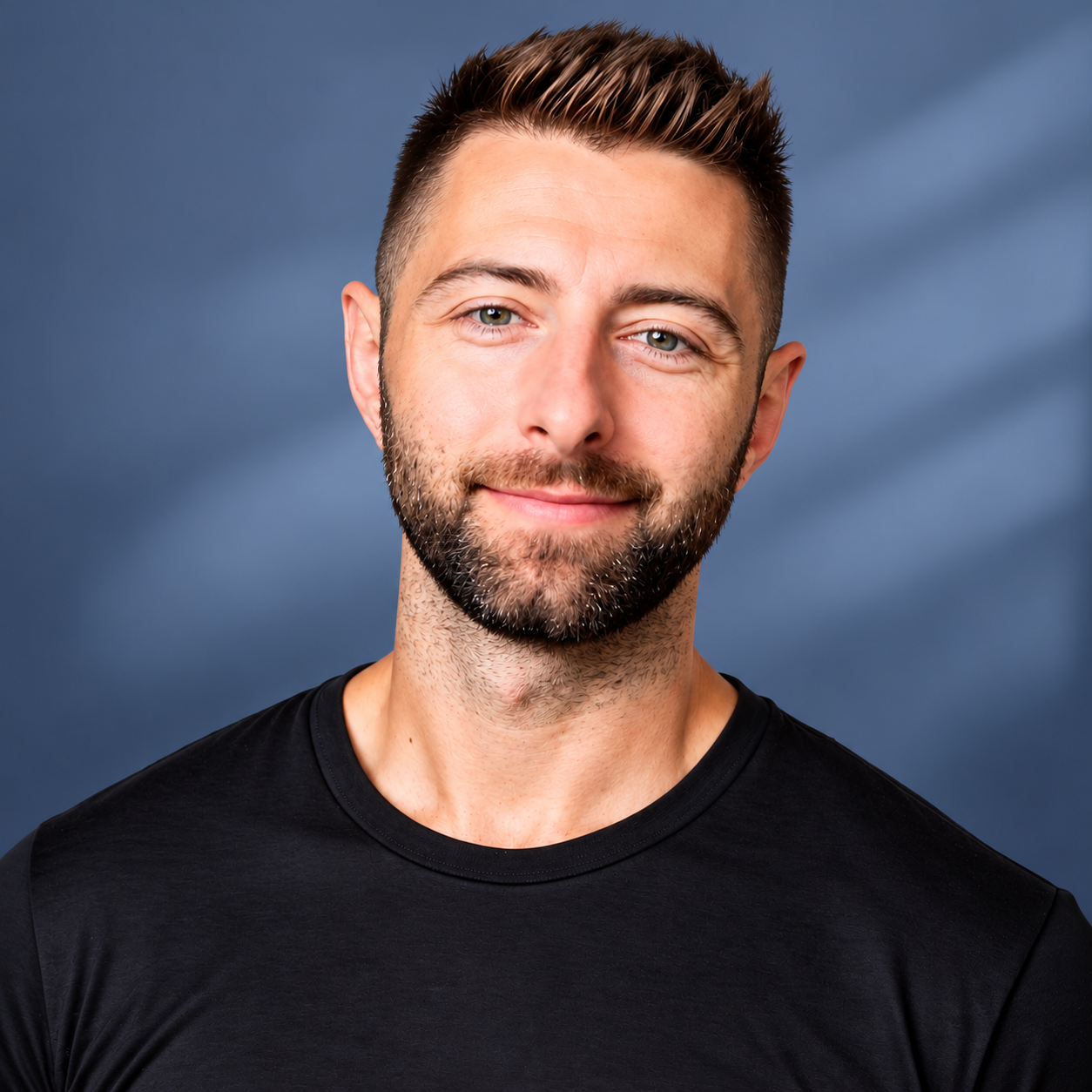 A smiling man with short brown hair and a beard, wearing a black t-shirt, standing against a dark cloudy sky background.