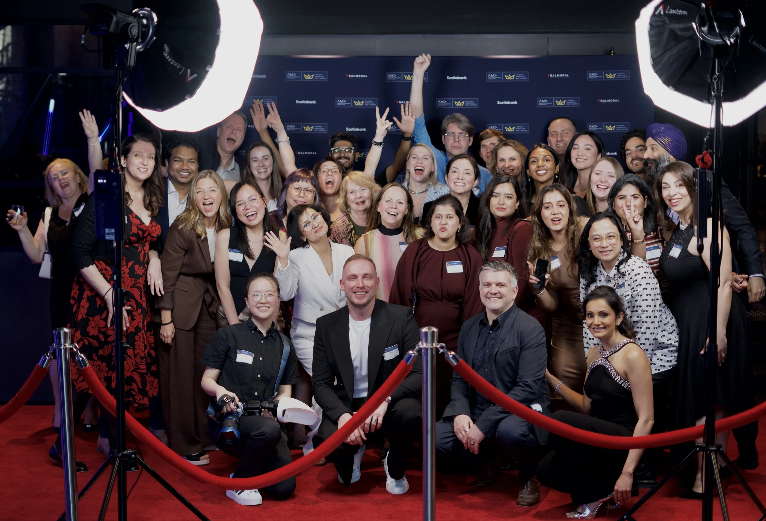 A large group of diverse people posing together behind a red rope barrier at an event. The backdrop features logos of Scotibank, AMD, and Balmoral, indicating a formal or corporate event setting. There are professional lighting setups overhead illuminating the group.