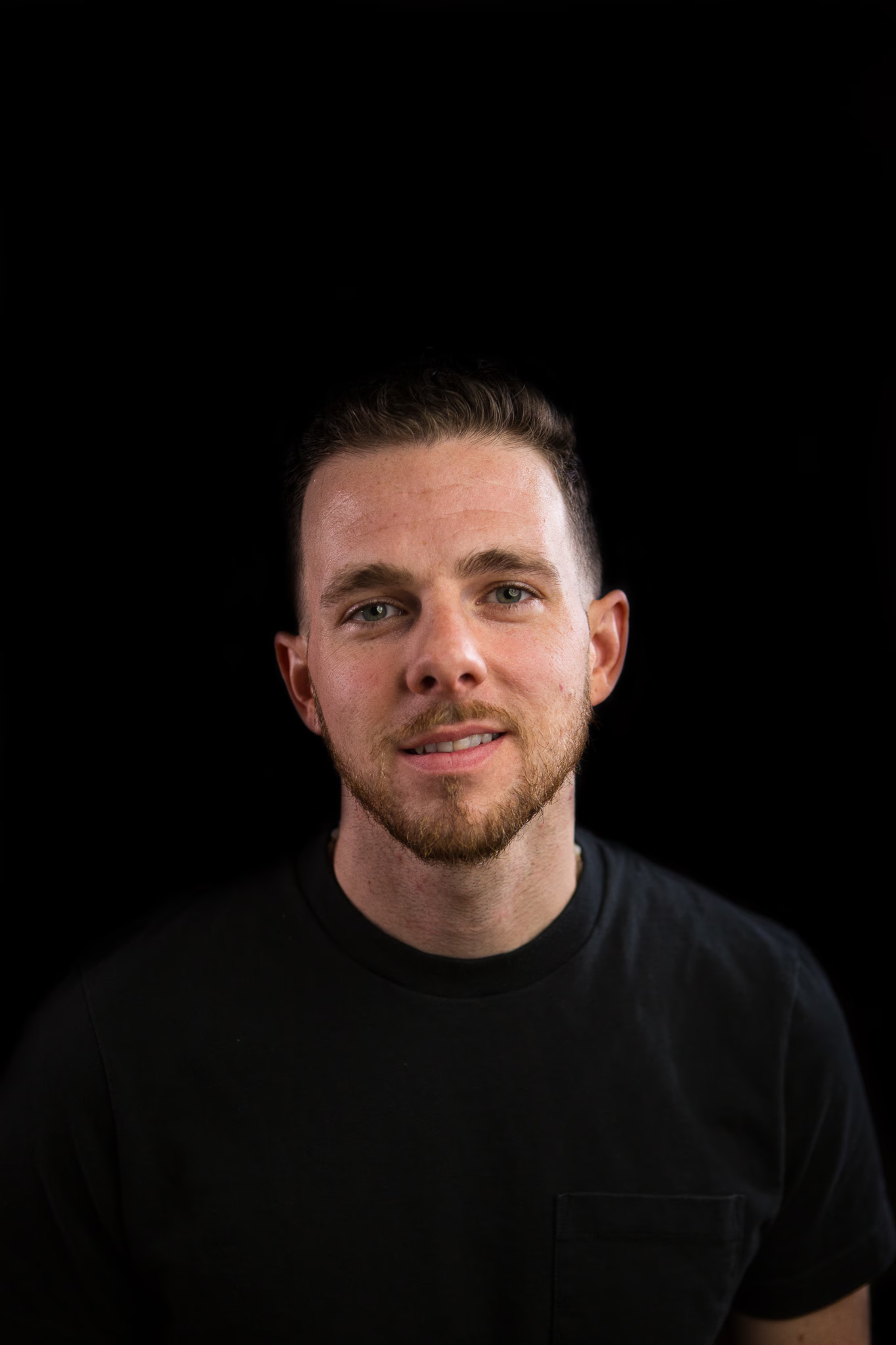 Close-up of a young man with short brown hair and a beard, wearing a black shirt, smiling slightly against a black background.