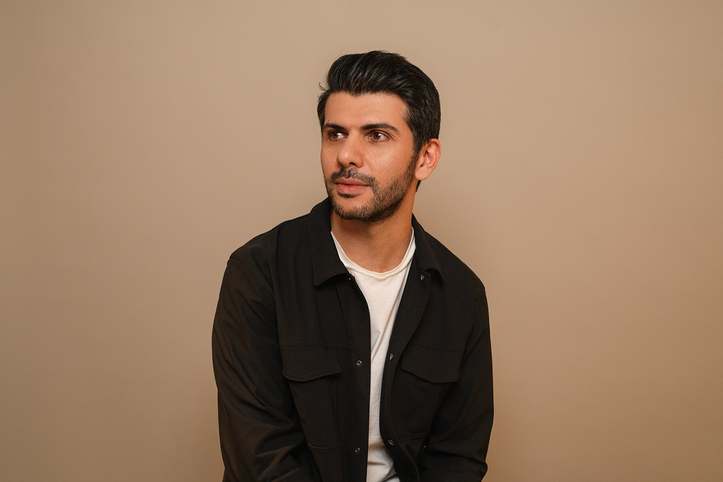 A man with dark hair and a beard, wearing a black jacket over a white shirt, looks to the side against a beige background.