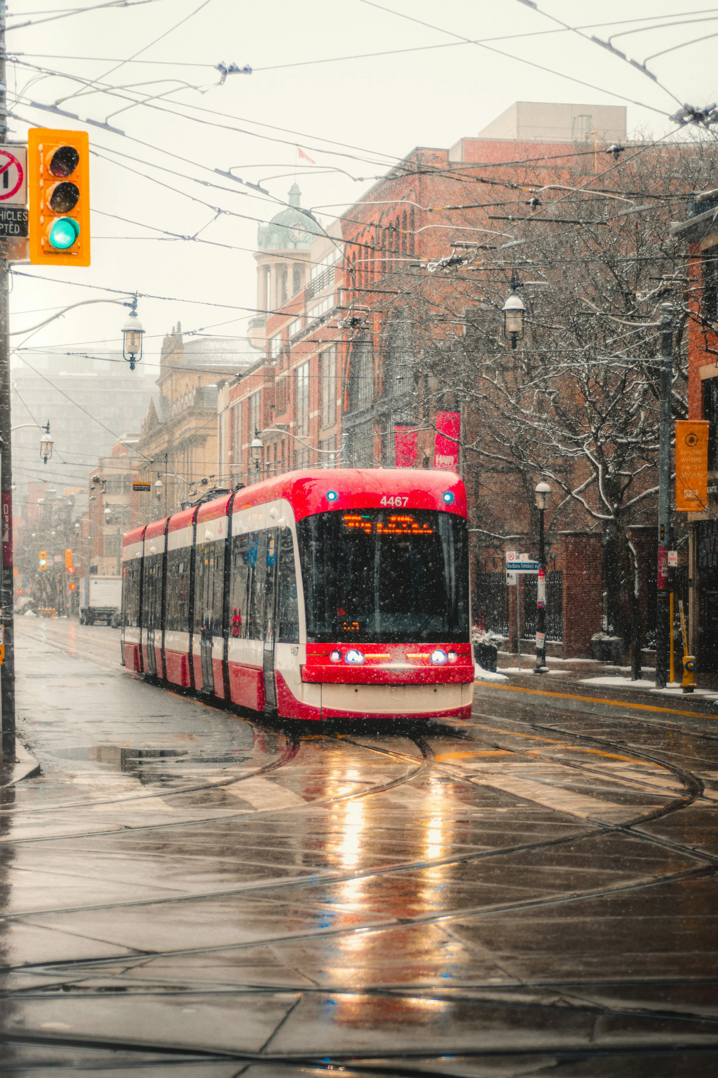 A red and white streetcar traveling on tracks in a city street during a rainy day with snow falling. Traffic lights and city buildings with red brick facades and a clock tower in the background.