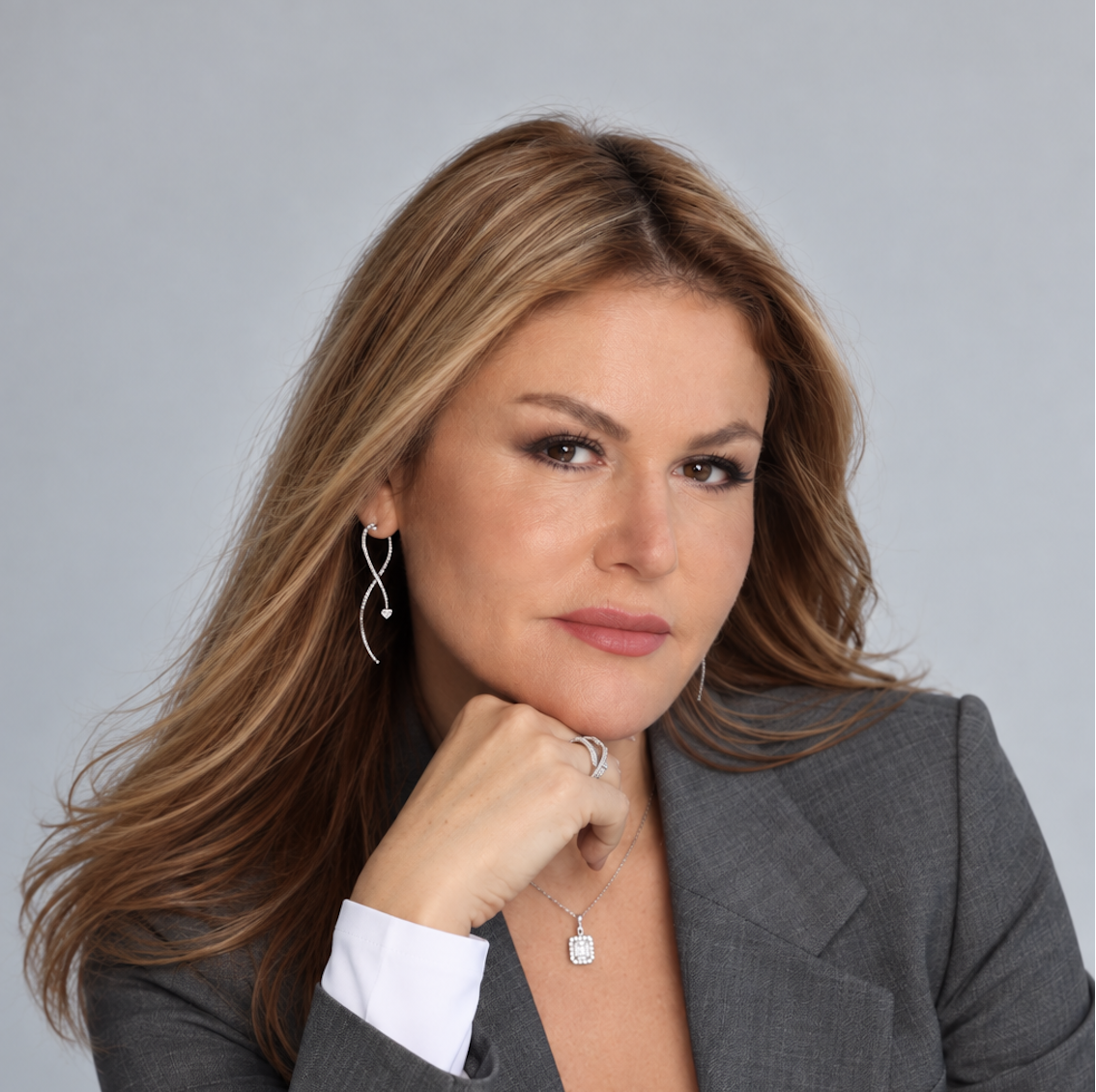 Close-up portrait of a woman with long reddish-brown hair, wearing a gray blazer, silver jewelry, and makeup, posing against a light gray background.