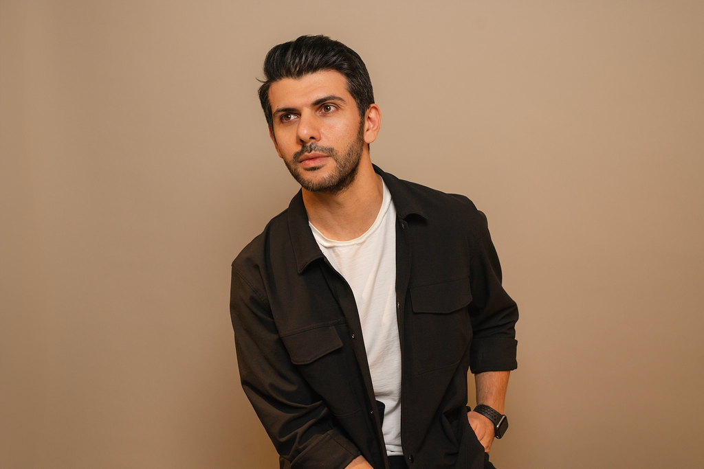 A young man with dark hair and facial hair, wearing a black shirt over a white t-shirt, standing against a plain, light-colored wall.
