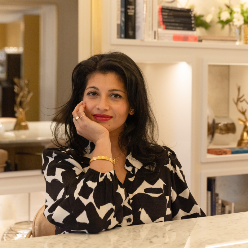 A woman with dark hair and red lipstick sitting at a table, resting her head on her hand, wearing a black and white patterned blouse, with a warm and inviting room background.