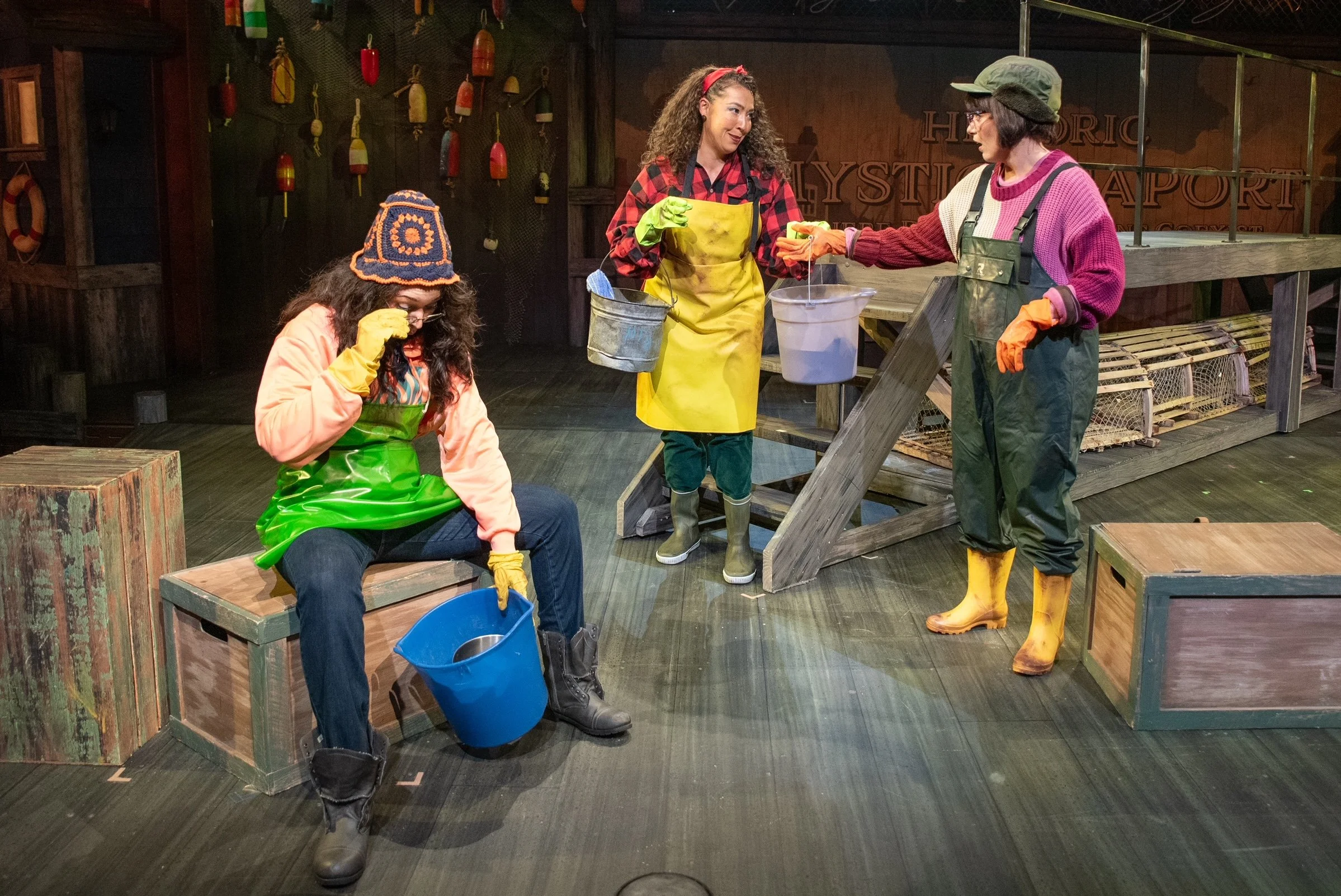 Three women on a stage set resembling a fishing or outdoor shop. Two women are standing, and one woman is sitting. They are dressed in colorful outdoor and fishing gear, with gloves and boots, engaged in a conversation or activity related to fishing or outdoor work. The background includes fishing floats and life preservers.
