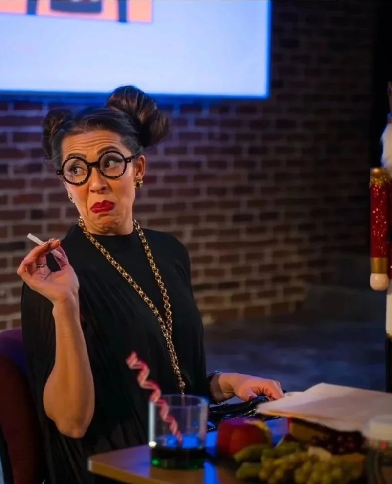 A woman with dark hair styled in two buns, wearing black glasses, a black dress, and gold jewelry, is sitting at a table holding a cigarette and making a facial expression of skepticism or disapproval. The table has papers, a drink with a red straw, and some snacks. The background features a brick wall and a television screen.