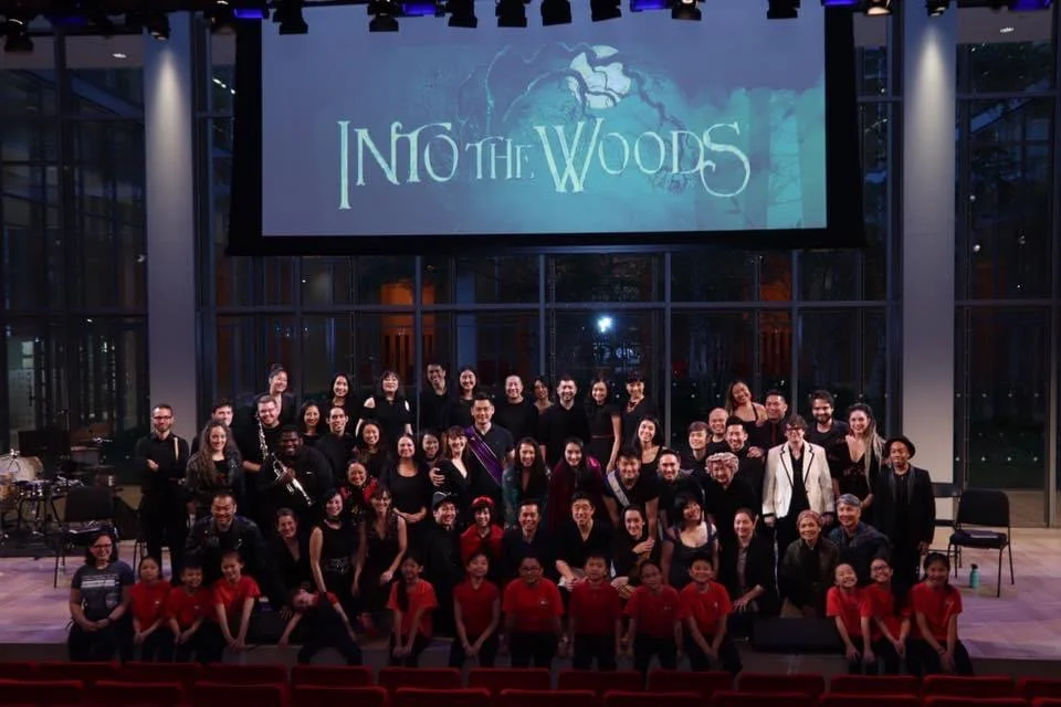 Group photo of children and adults on a stage with a large screen displaying 'Into the Woods' in the background.