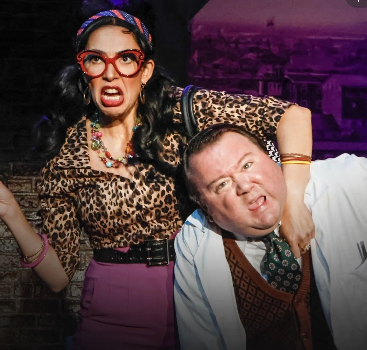 A woman with dark curly hair, red glasses, and colorful jewelry is aggressively holding a man in a white shirt, who is grimacing and sticking out his tongue. They are on a stage with a dark background.