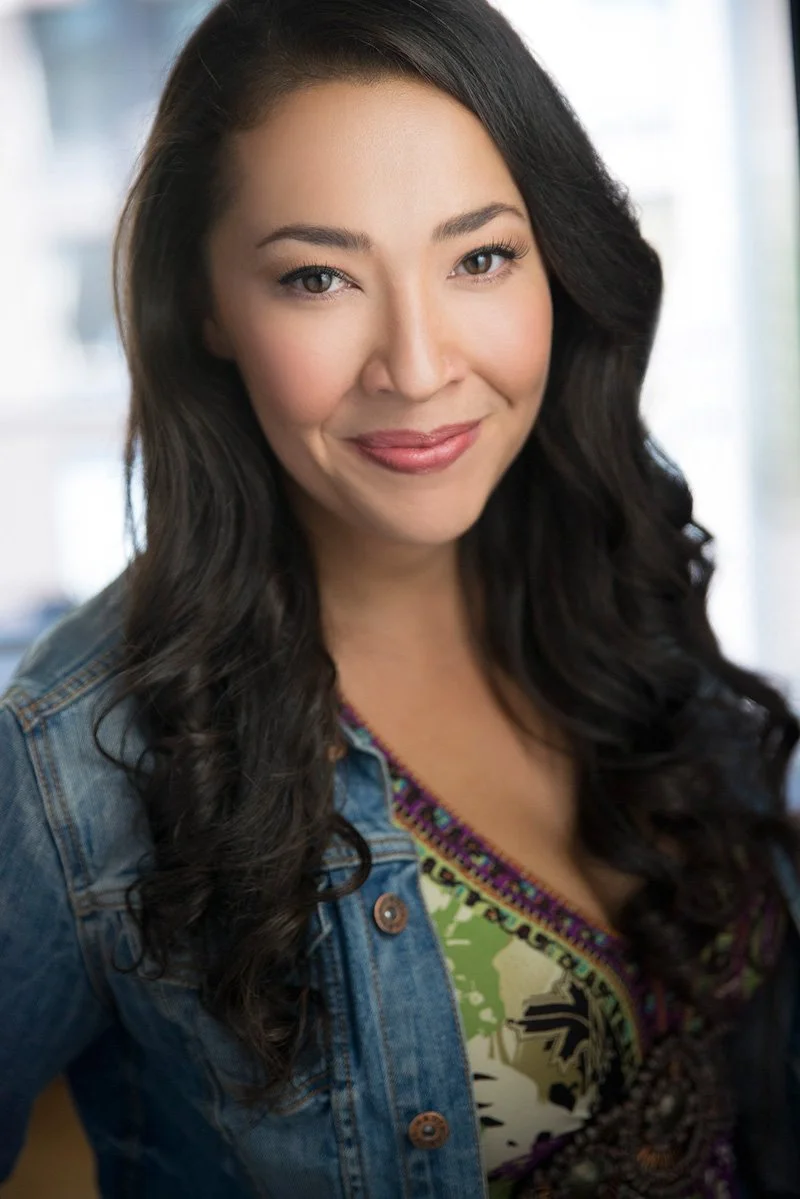 A woman with long, curly black hair, wearing a denim jacket and a colorful patterned top, smiling at the camera with an indoor background.