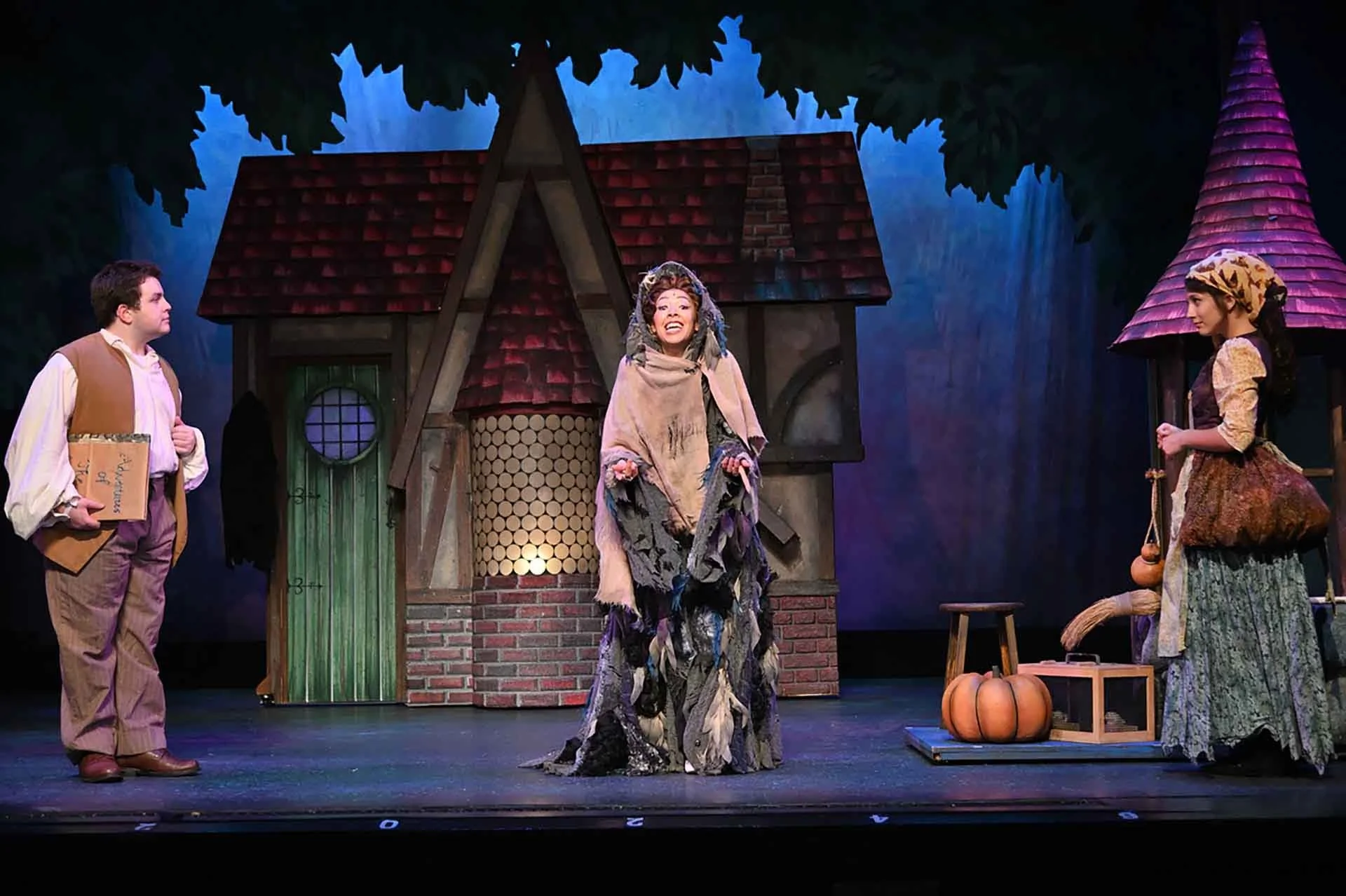 Actors performing a scene on stage dressed as characters from a fairy tale. The set includes a small house with a red shingled roof, a green door, and a chimney. One woman in ornate costume is center stage, smiling, with two women and a man in period costumes on either side, surrounded by props like pumpkins and a stool.