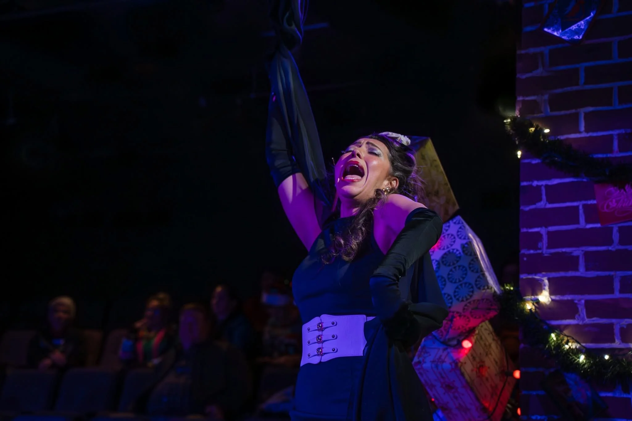 A woman performing on stage with her arm raised, wearing a black dress and white corset, singing passionately in front of a crowd in a dimly lit setting with purple and blue lighting.
