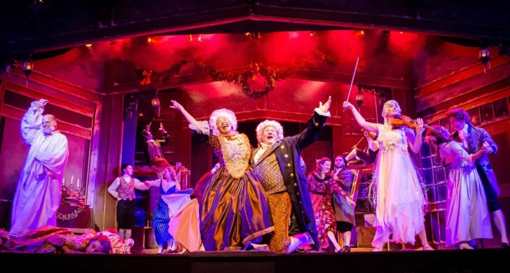 The stage of a theatrical performance with actors dressed in period costumes, celebrating or performing a lively scene with music and dance near Christmas decorations and warm lighting.
