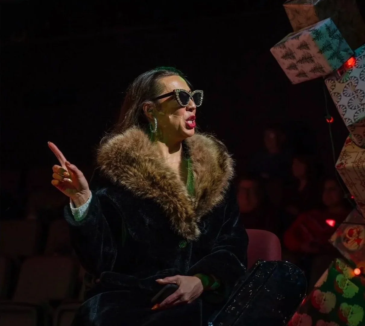 A woman wearing sunglasses, a fur-collared coat, and earrings, sitting in a dark room and gesturing with her hand.