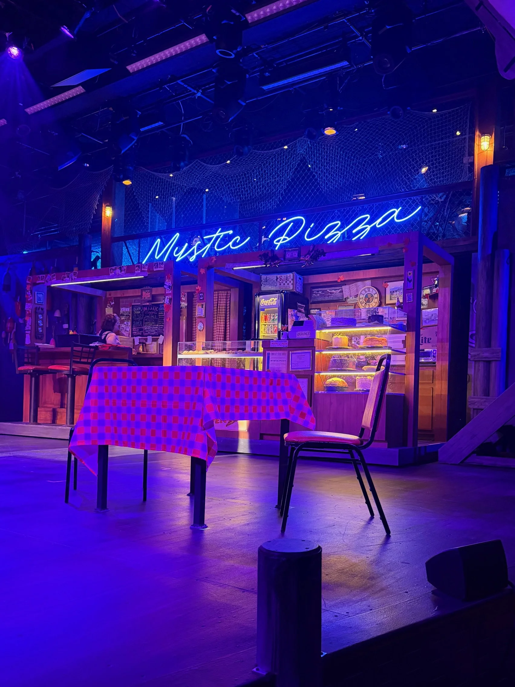 Stage with blue neon sign reading 'Mystic Pizza', tables, chairs, and a pink checkered tablecloth in the foreground, dim lighting, and a food counter in the background.