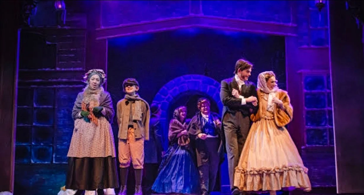 Actors performing on stage in a theatrical production set in a historical period, featuring seven actors in period costumes, with a dark background and purple lighting.