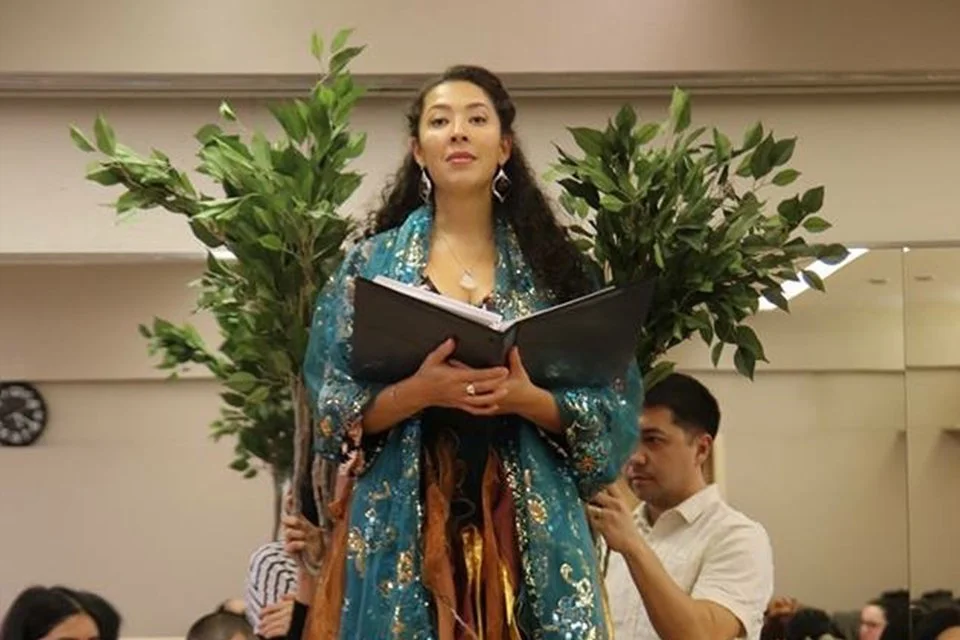 A woman holding a book, standing outdoors, with trees behind her and people around.