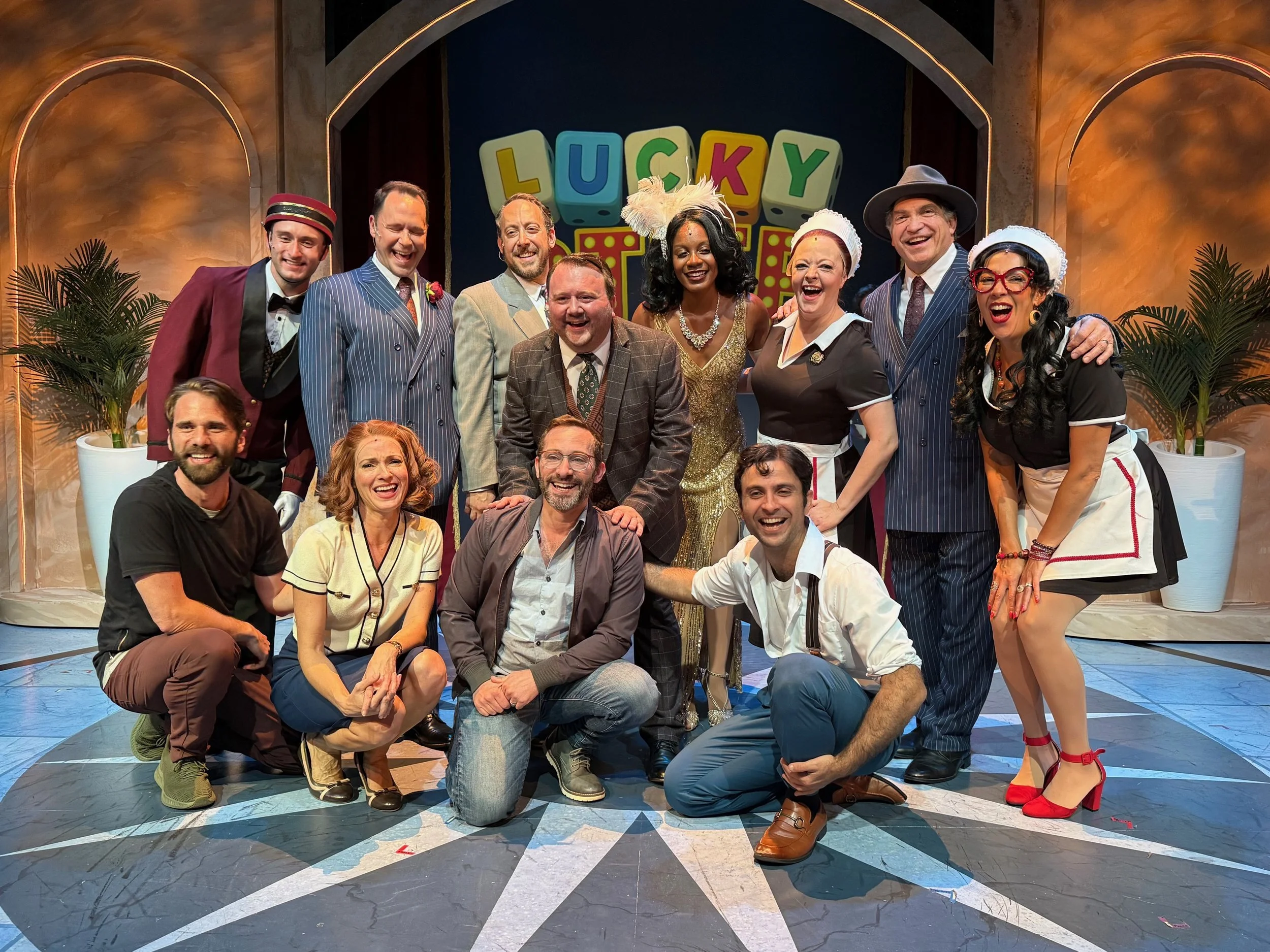 Group of actors in colorful costumes on stage, smiling and posing for a photo, with a backdrop that says 'Lucky' in large letters.