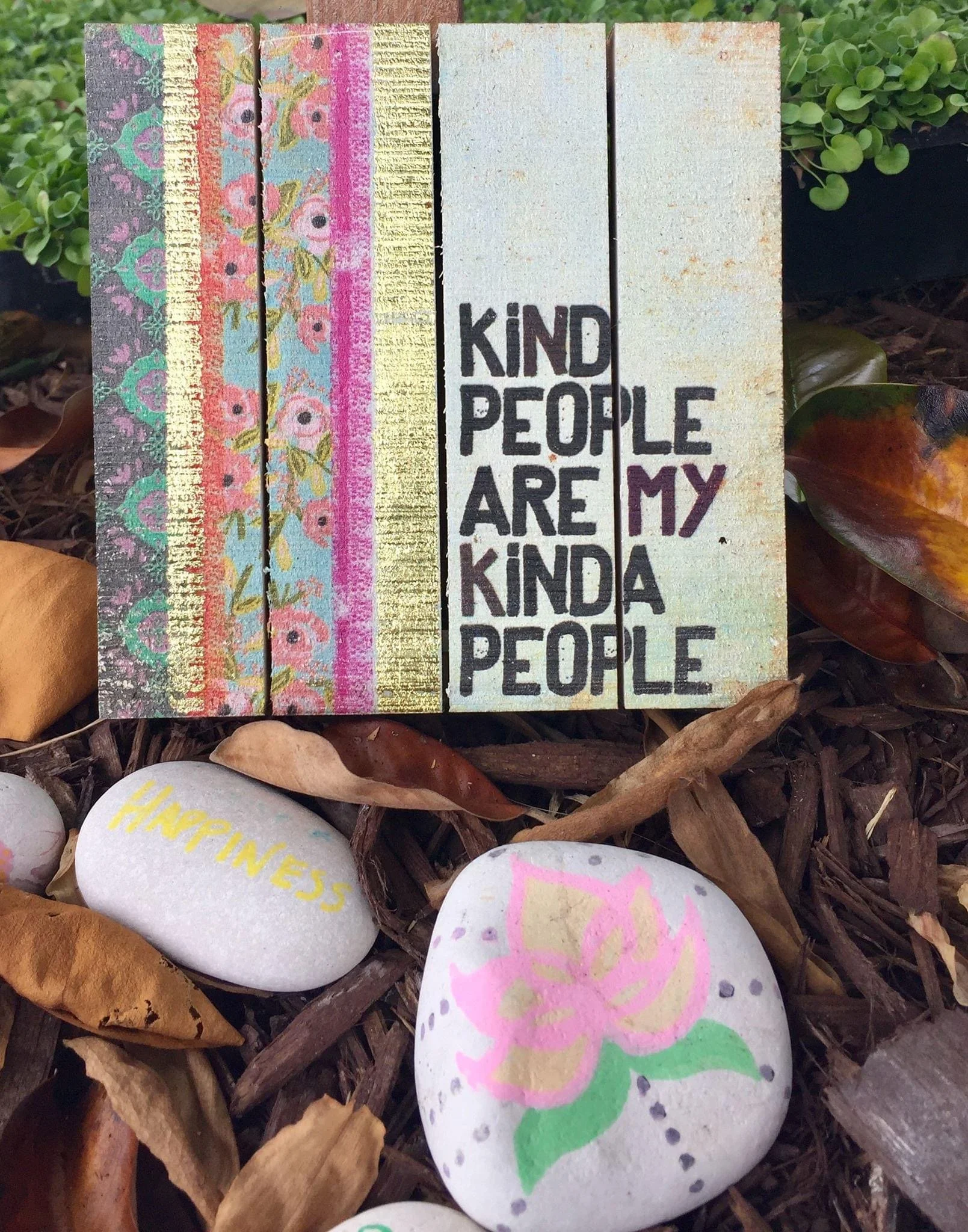 Decorative painted rocks with messages like 'kind people are my kinda people,' surrounded by leaves and mulch.