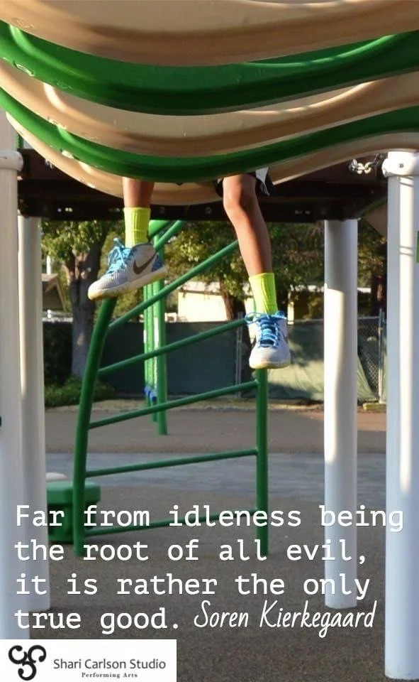 Child hanging upside down from a playground structure, legs extended downward, wearing blue and white sneakers with neon yellow socks. A quote overlaid at the bottom of the image reads: 'Far from idleness being the root of all evil, it is rather the only true good. Soren Kierkegaard.'