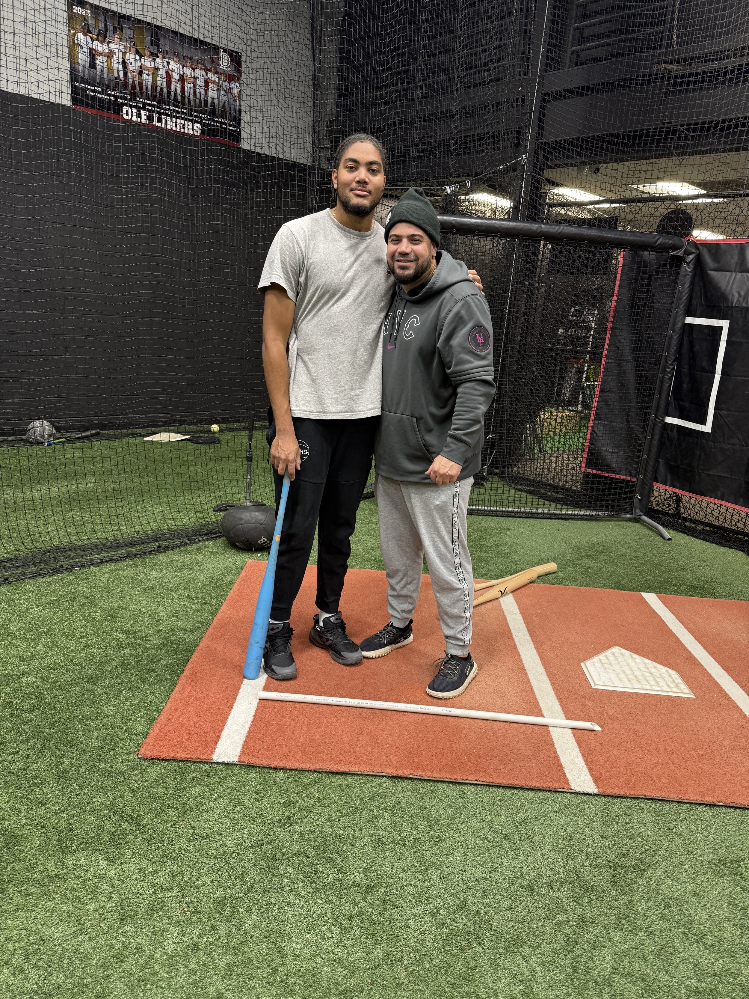 Two men are standing on a small baseball or softball field, smiling for a photo. The taller man on the left is holding a baseball bat, and both are dressed in casual athletic clothing. They are standing on an orange mat with base lines visible, with a baseball infield setup around them. The background features a black safety net, some sports equipment, and a poster of baseball players.