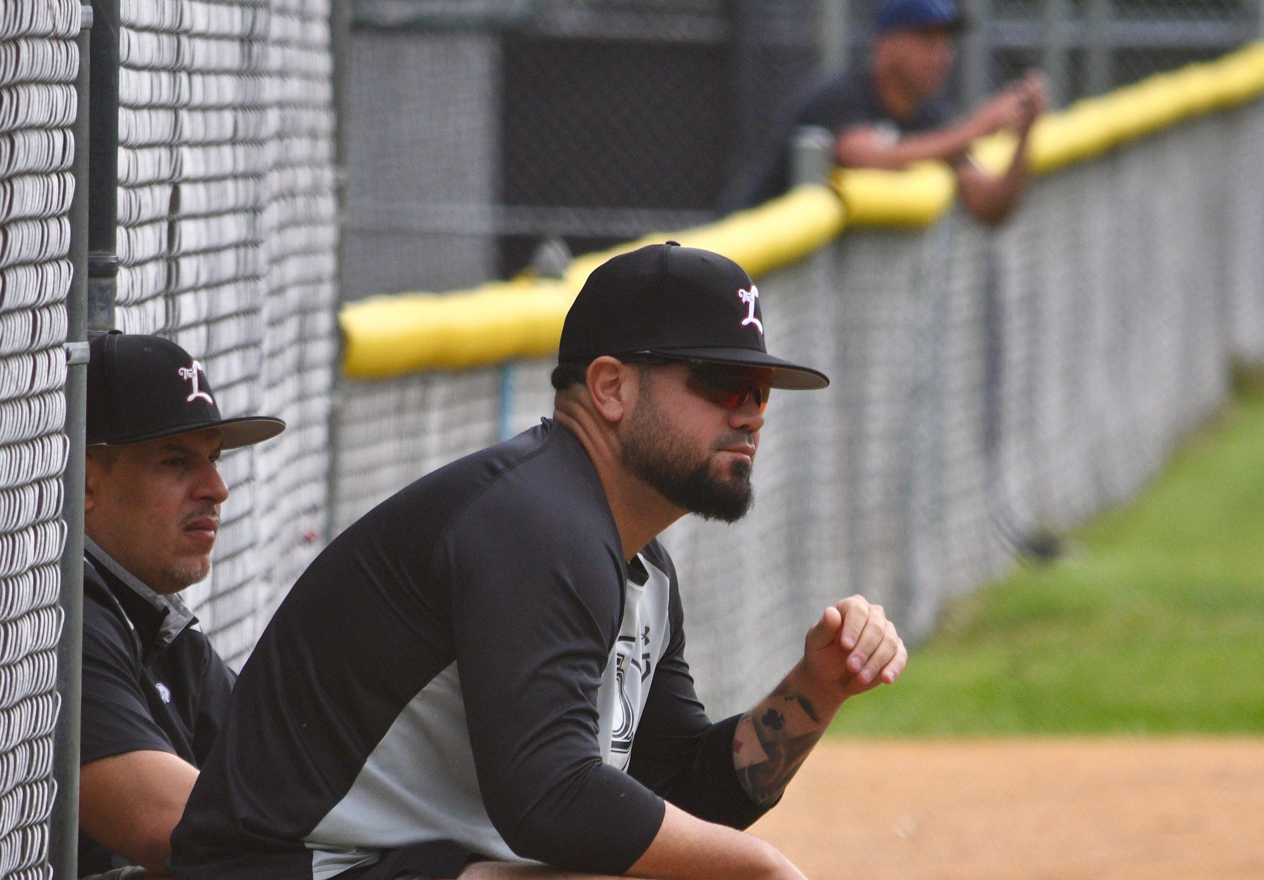 Two baseball players sitting in the dugout, wearing black and white uniforms and black caps with a red and white logo. One player is looking forward, the other is leaning on the yellow railing, with both appearing to be in a moment of reflection or concentration.