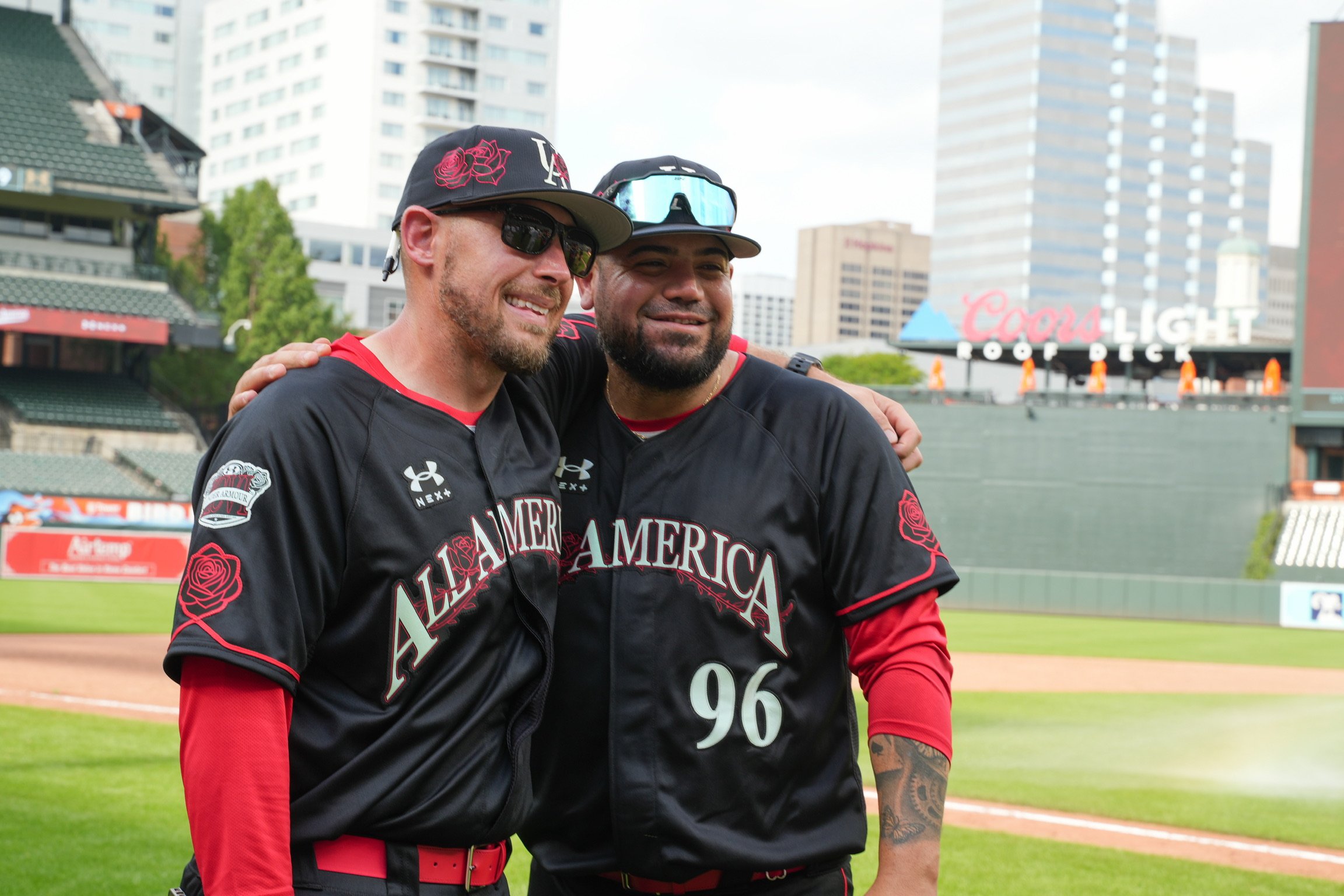 Two baseball players standing close together on a baseball field, smiling, with city buildings in the background. One player is wearing black with red accents, sunglasses, and a cap, while the other has a beard, a cap, and sunglasses on his head.
