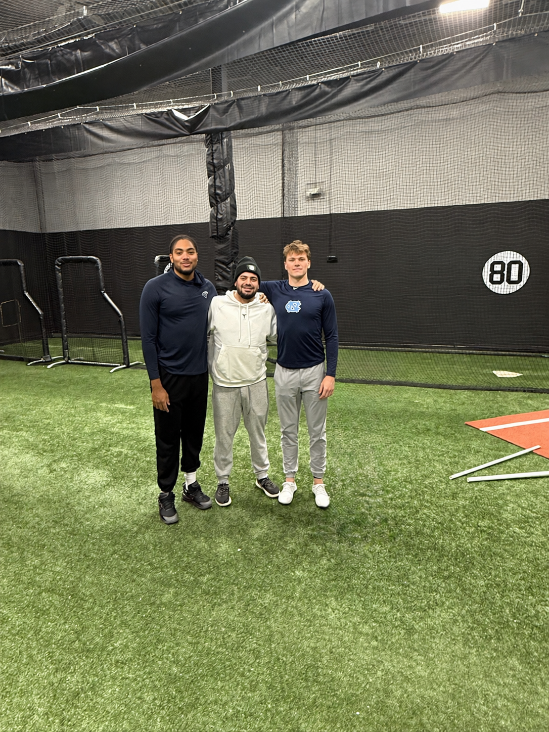 Three young men standing together in an indoor sports facility with green artificial turf and black mesh walls, smiling at the camera.