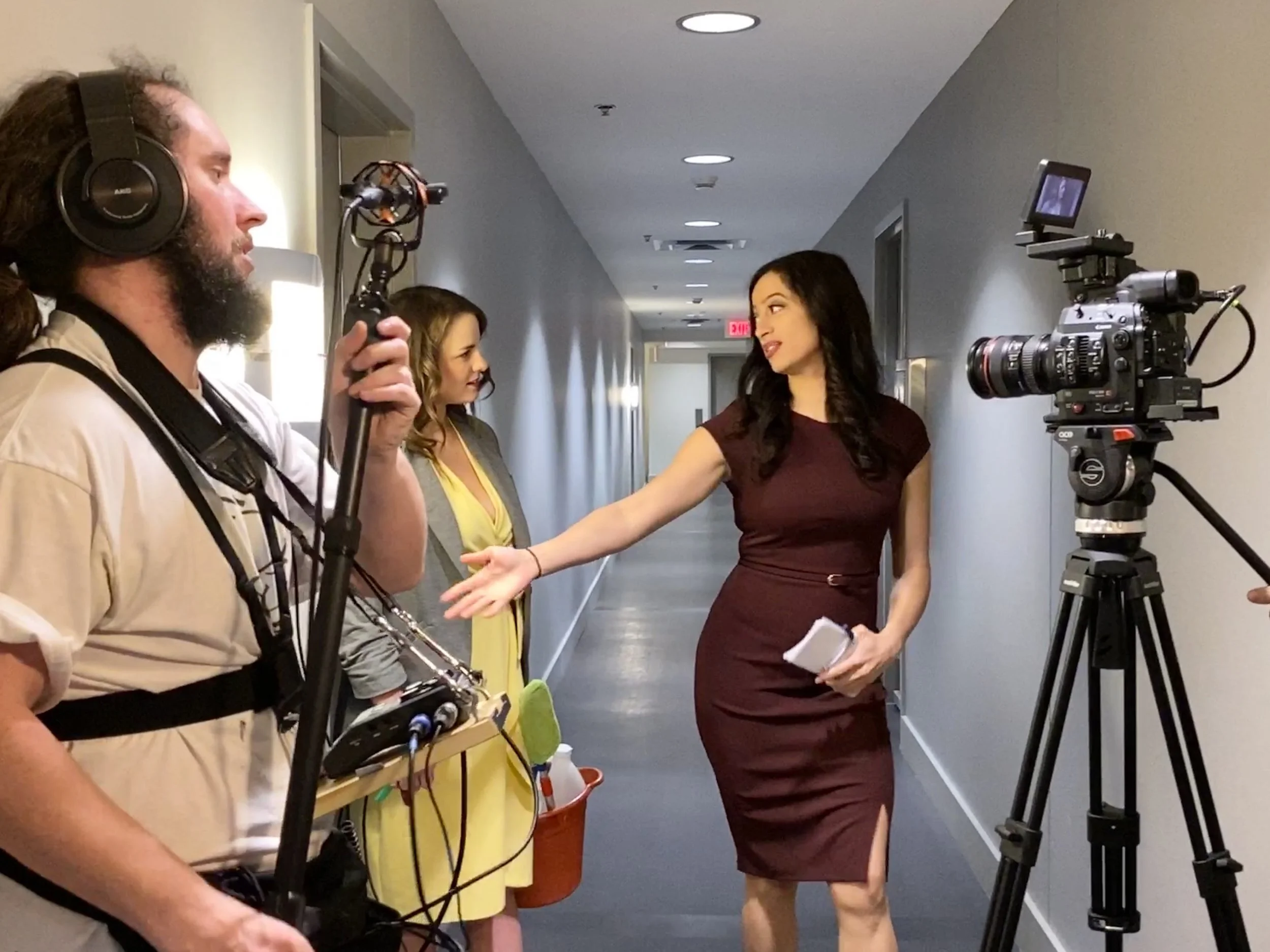A woman with dark hair in a burgundy dress is speaking to a camera crew, gesturing with her right hand. A man with a beard, headphones, and beige clothing, holding a camera stabilizer, is on the left. Two women are in the background, standing in a hallway with white walls and ceiling lights, one with curly hair and a gray blazer, the other in gray and yellow. A professional video camera mounted on a tripod is on the right.