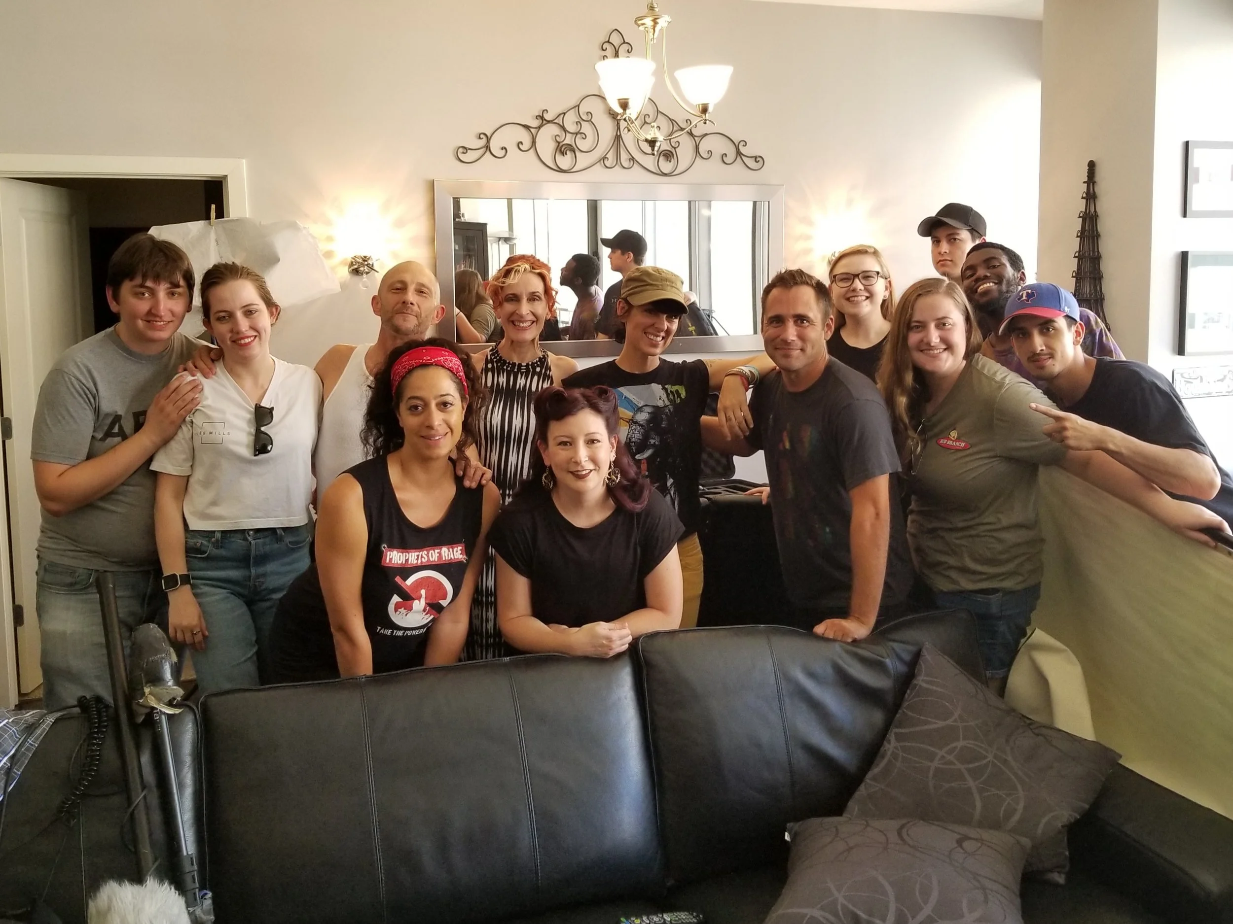 A group of about fifteen people, diverse in age and ethnicity, smiling and posing together in a living room or common area. They are standing in front of a mirror and a window, with some making peace signs or pointing at each other. The environment appears casual and friendly, with chairs and a television remote visible in the foreground.