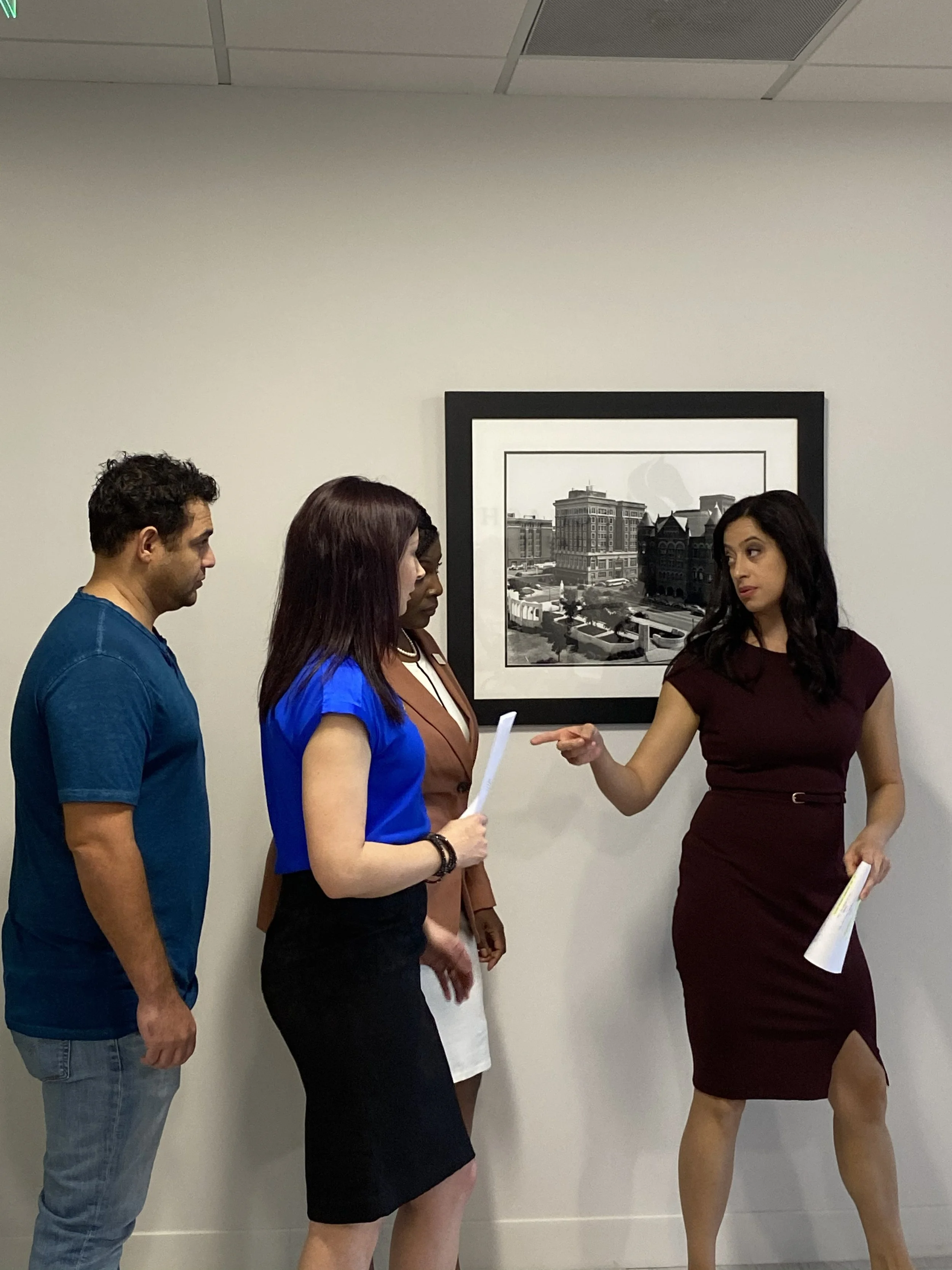 Four women and one man engaged in conversation in an indoor setting, standing in front of a wall with framed black-and-white cityscape photograph.