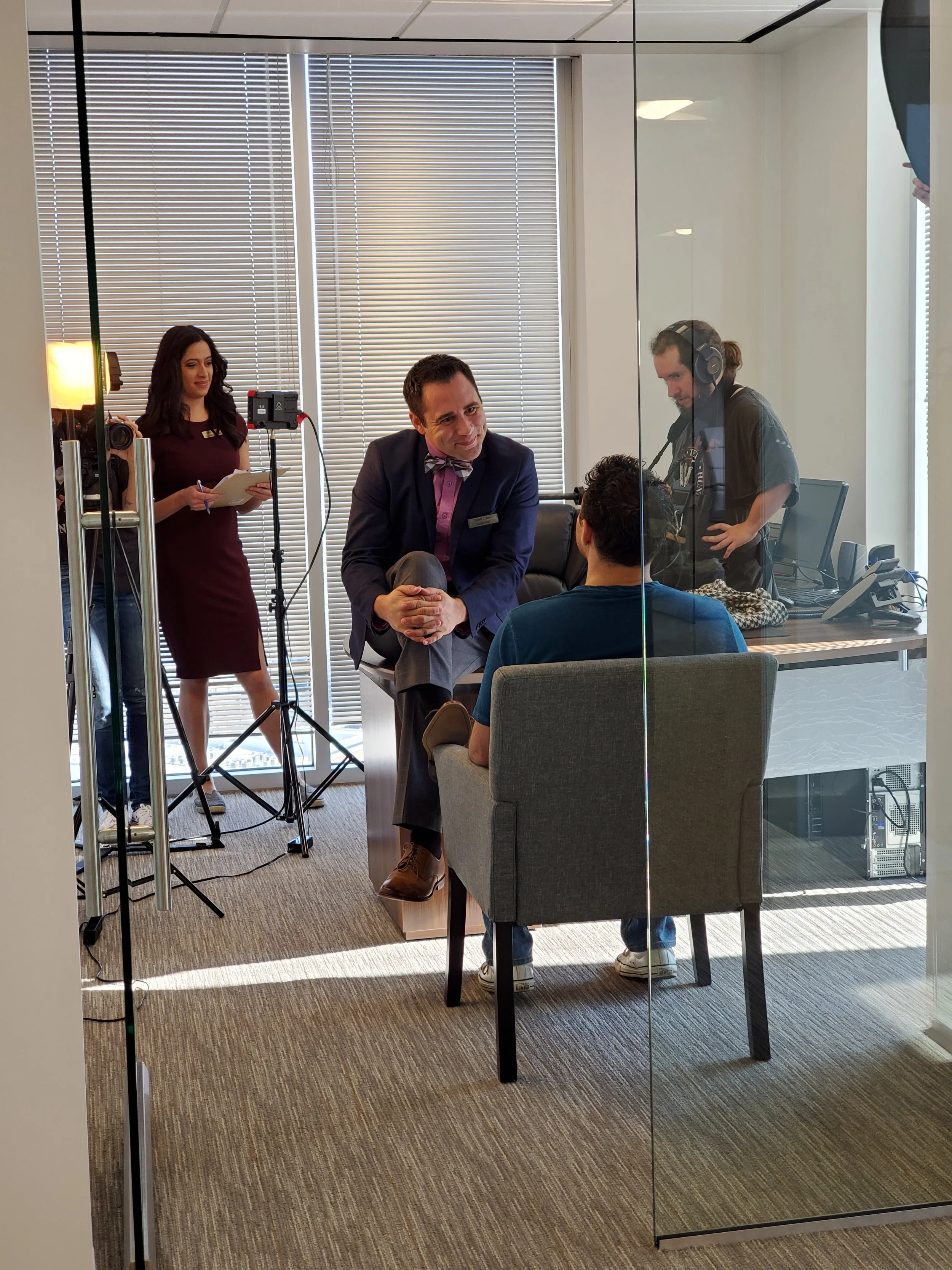 View through a glass wall showing a conference room with five people. One woman is standing with a notepad, and a man in a suit with a bow tie is sitting and talking to another man seated with his back to the camera. A person with headphones is working at a desk with computer equipment. The room has window blinds and carpeted flooring.