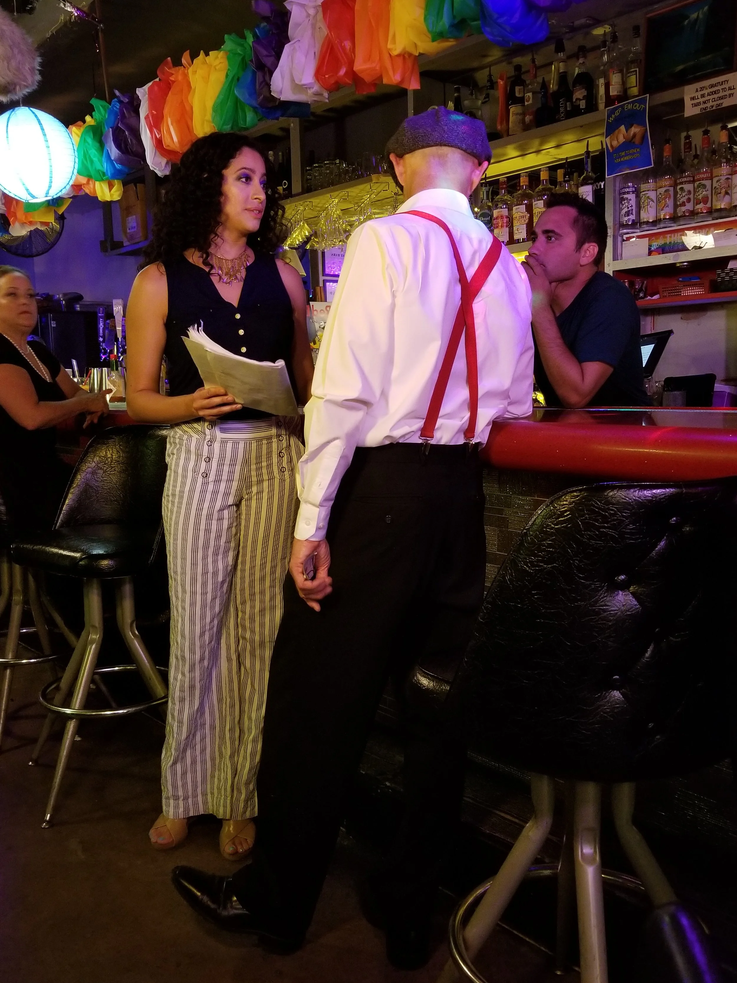 A woman and two men are in a bar, engaged in conversation. The woman is holding a menu, and the man facing away is wearing a beret, white shirt, red suspenders, and dark pants. There is another woman in the background sitting at the bar. The bar is decorated with colorful paper decorations and has various bottles of alcohol on the shelves.