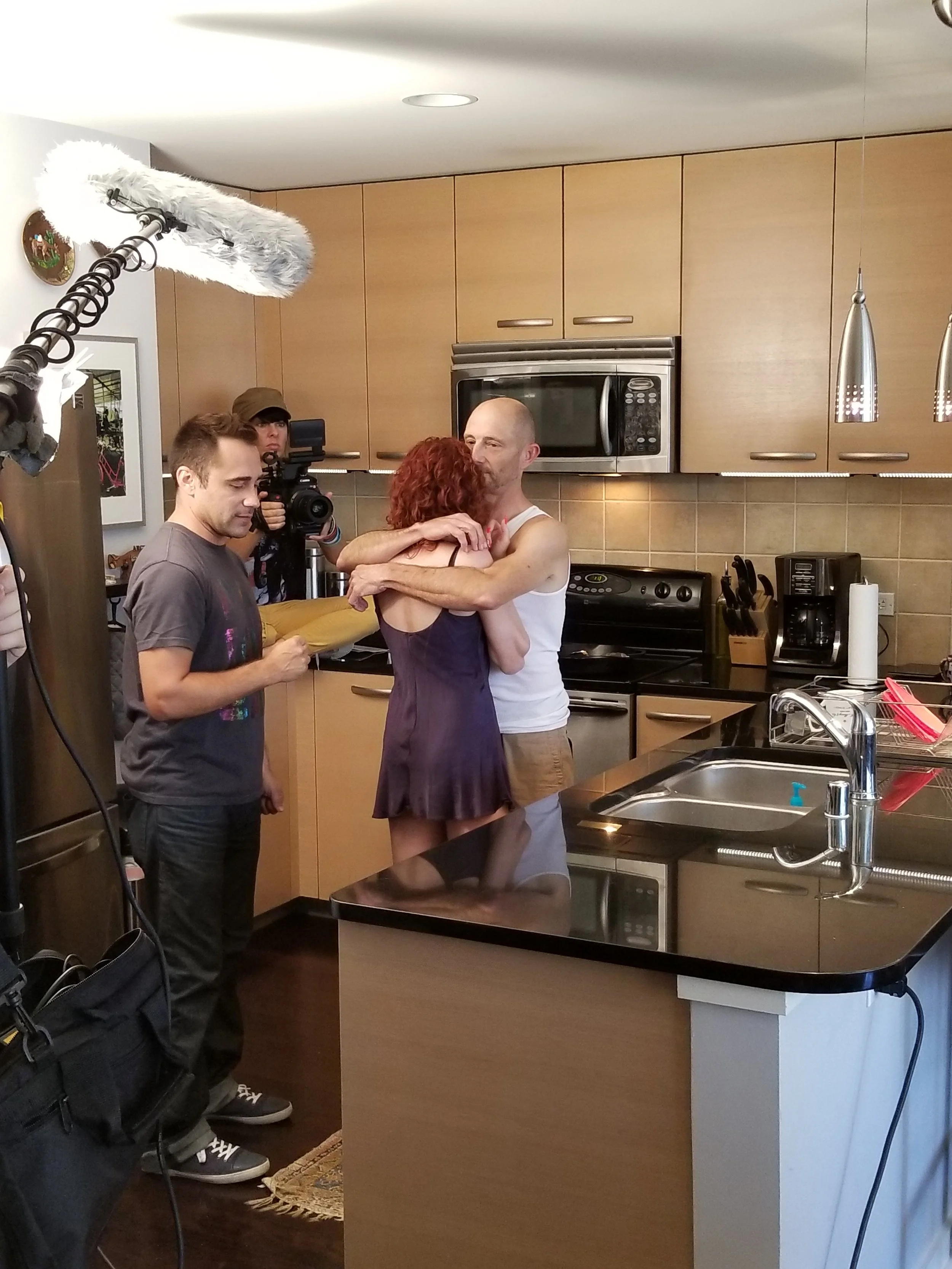 A man and woman hug in a kitchen, surrounded by crew members and a film camera, with filming equipment in the foreground.