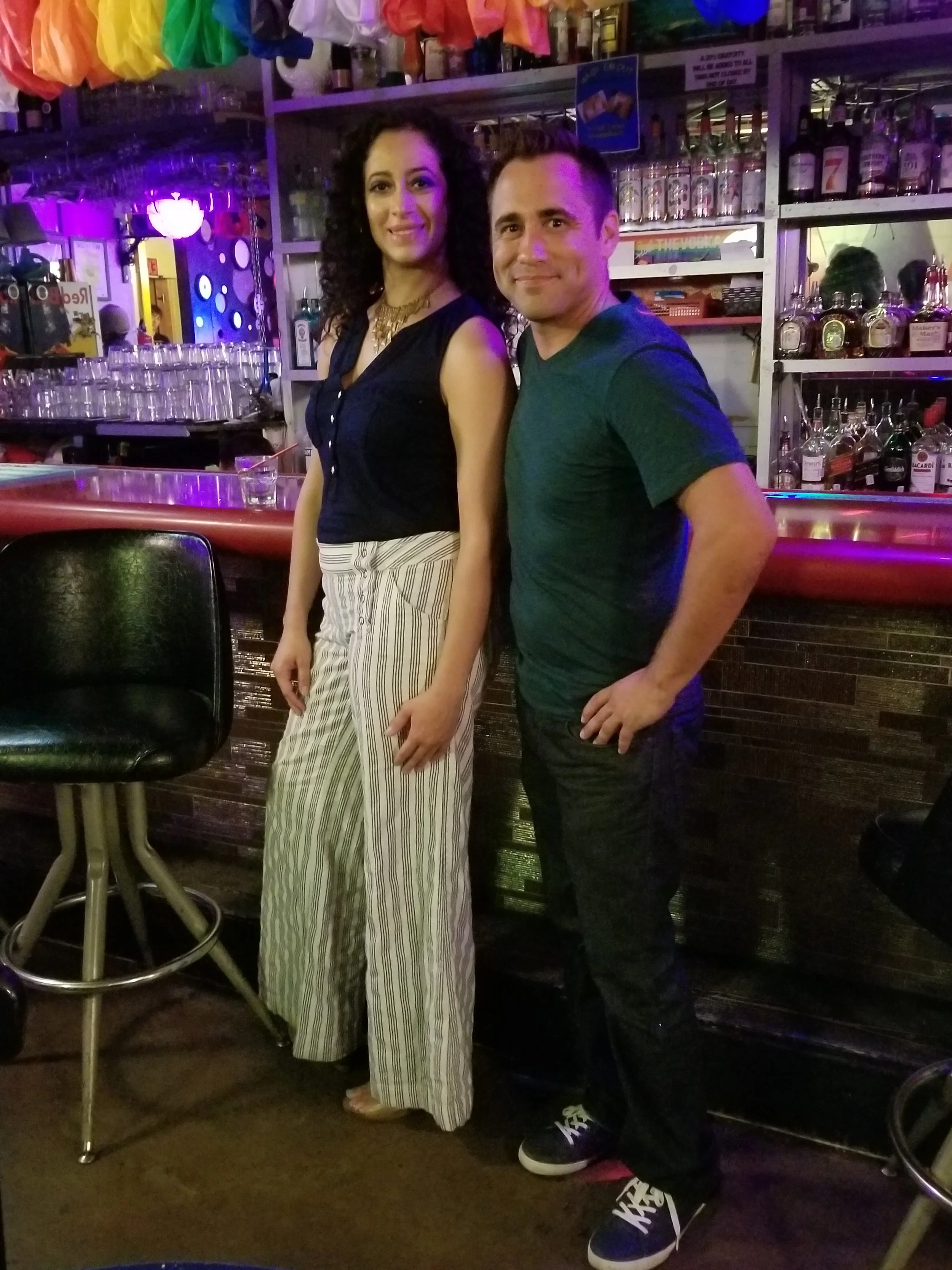 A woman and a man standing in a bar, smiling at the camera. The woman has dark, curly hair and is wearing a navy top with gold jewelry and striped pants. The man has short hair, wearing a dark green t-shirt and dark pants. The background features bar shelves with liquor bottles and colorful decorations, with a dark, cozy atmosphere.