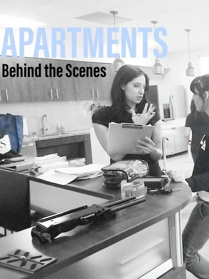 A woman is standing behind a kitchen counter, holding a clipboard, and gesturing with her hand as if explaining something. There are various items on the counter, including papers, a stapler, and some office supplies. The background features kitchen cabinets, a sink, and modern pendant lights.