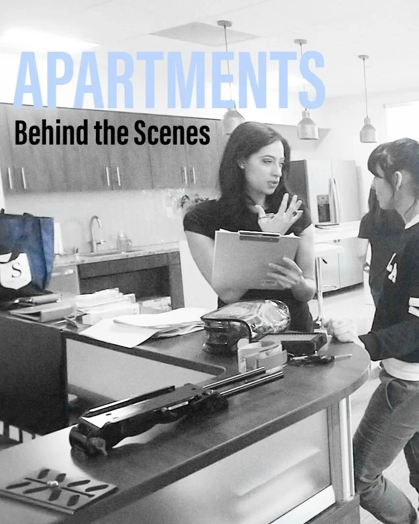 Two women in a modern kitchen, one holding papers and gesturing, the other listening, with a countertop filled with office supplies and a gun on the counter. Overhead lights and wooden cabinets are visible.