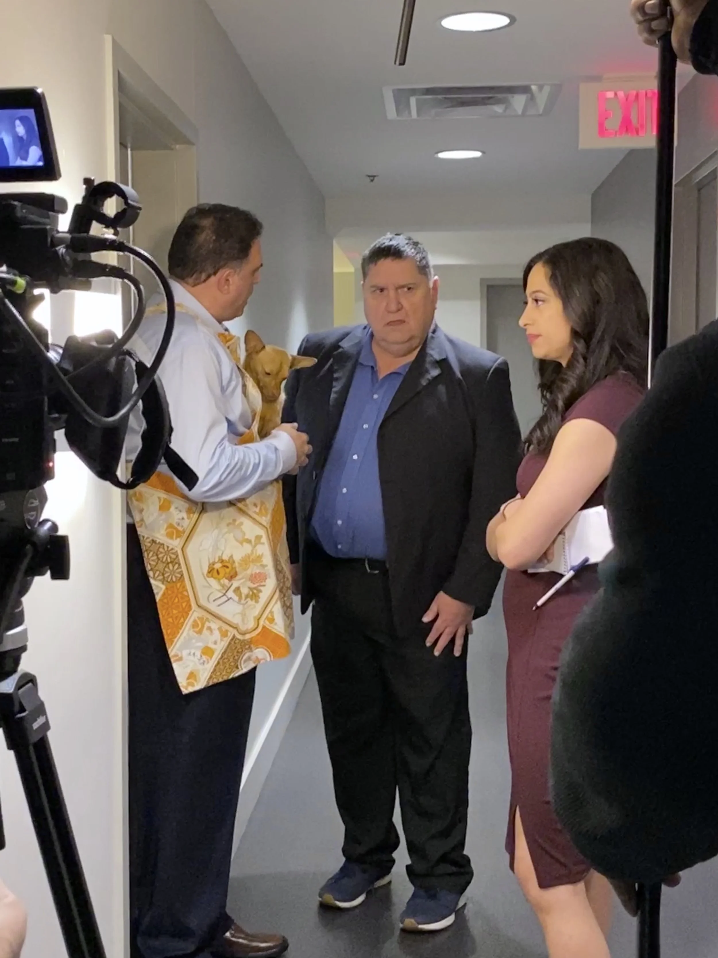 Group of four people standing in a hallway with one holding a small dog, engaged in conversation, with a camera on a tripod recording them.