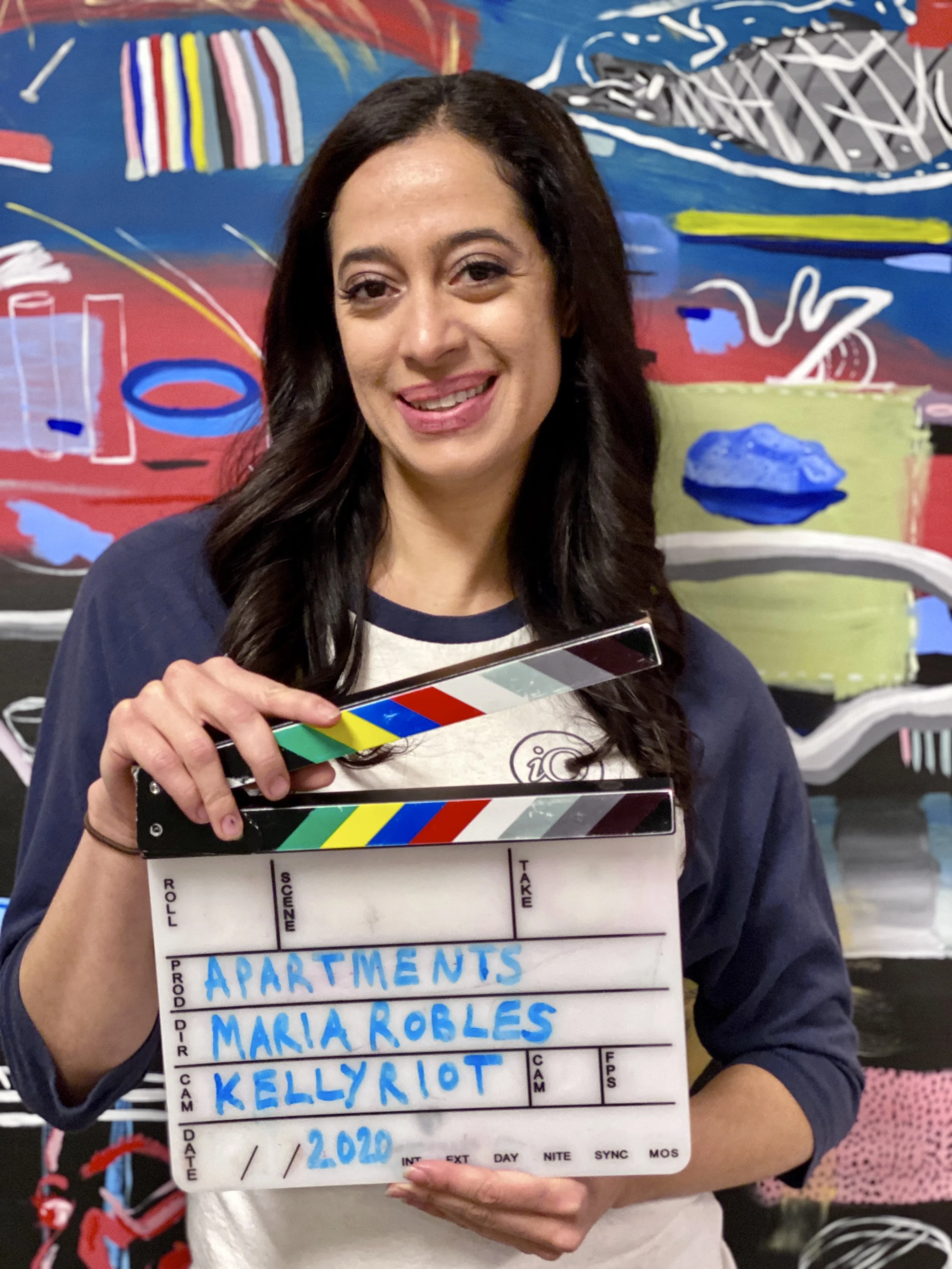 A woman holding a clapperboard with blue handwritten names including Maria Robles, Kelly Eliot, and others. She is in front of a colorful abstract painting.